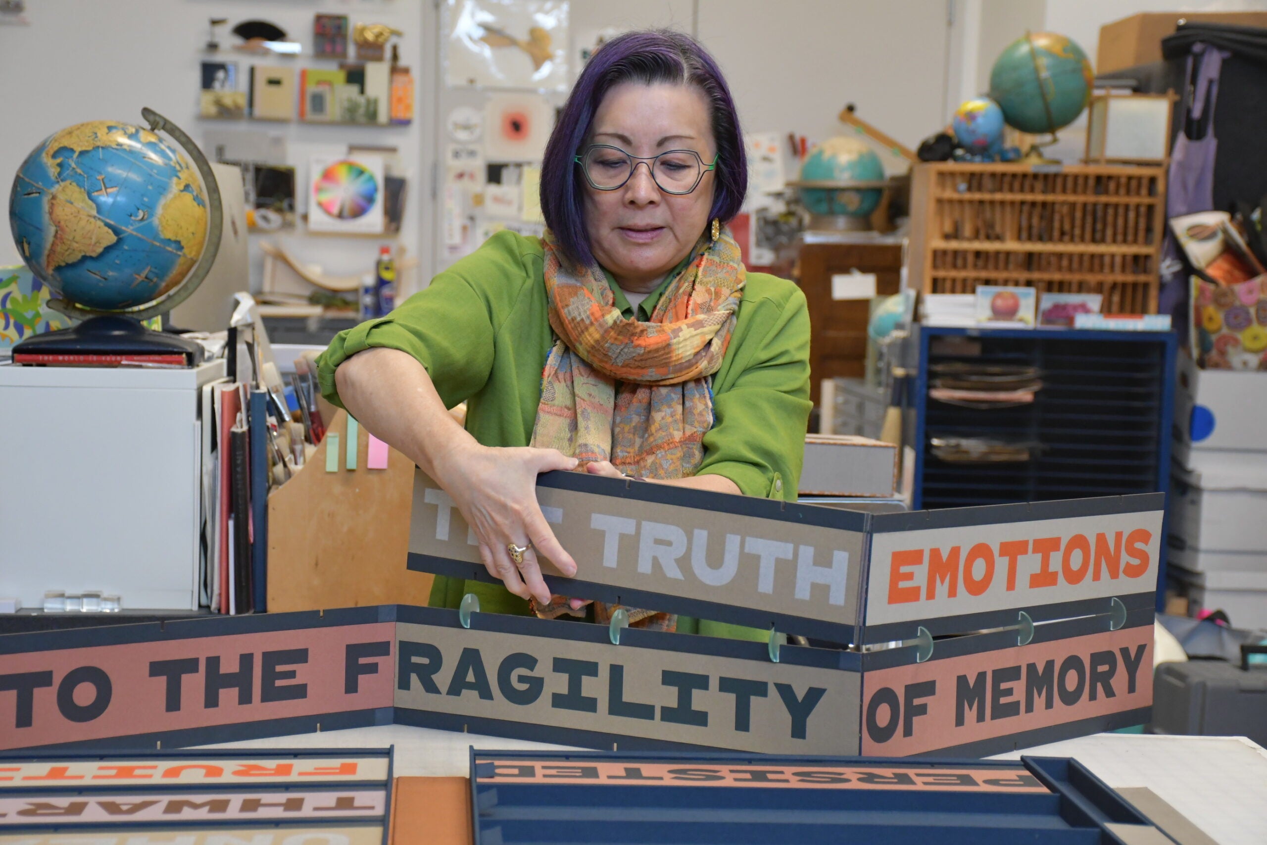 A woman with purple hair arranges colorful boxes labeled with words like TRUTH, EMOTIONS, and FRAGILITY OF MEMORY in a classroom filled with educational materials.