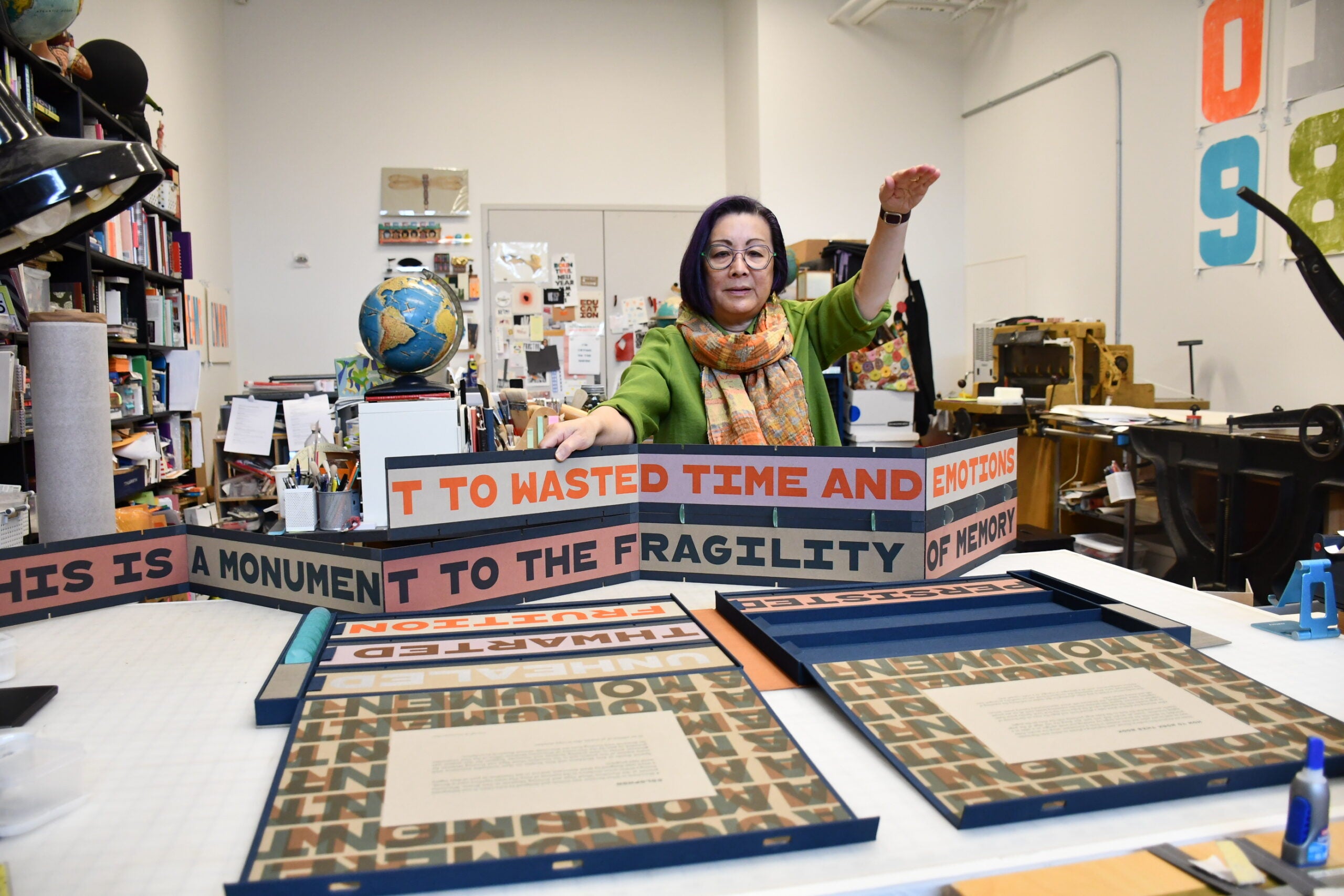 A woman stands in a studio, holding up a colorful, accordion-style art piece with bold text. Books, a globe, and artwork are visible around her on tables and shelves.