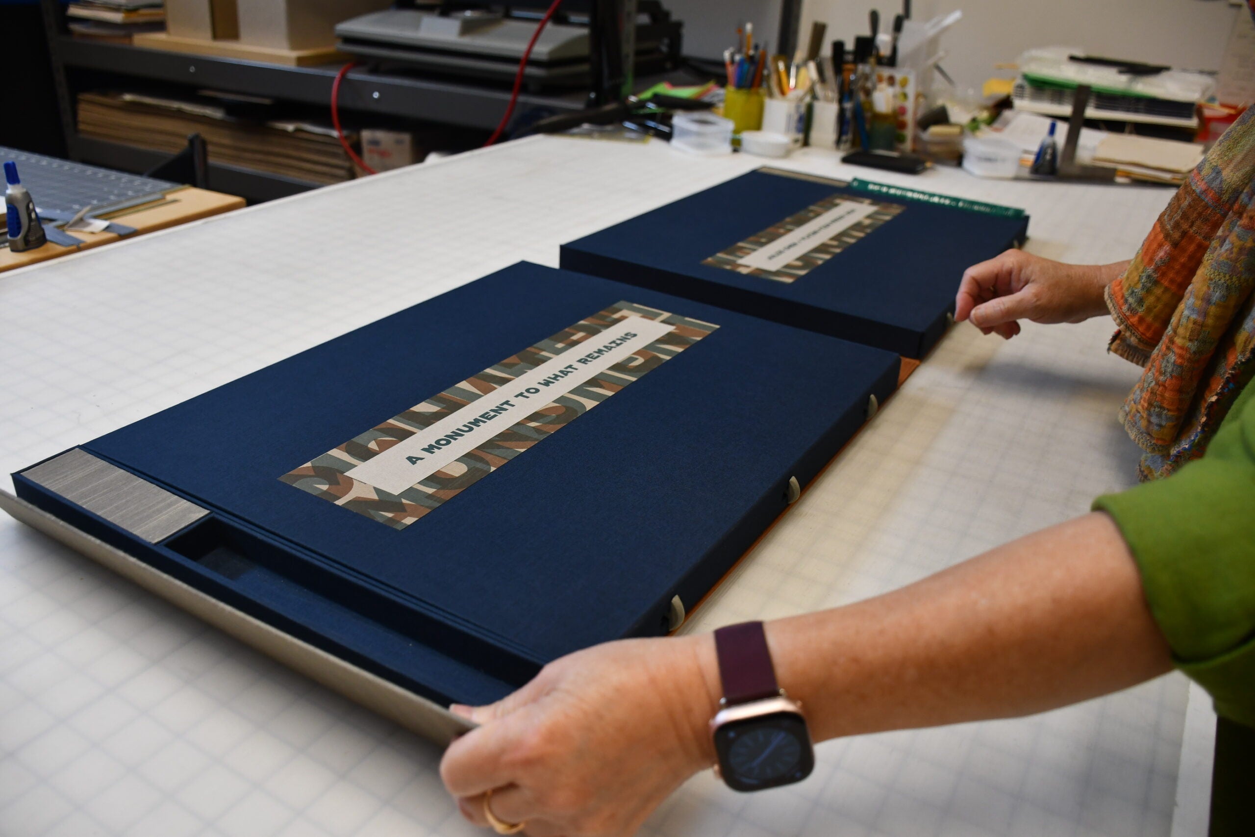 A person opens a large navy-blue portfolio labeled A MONUMENT TO MANY HEROES on a worktable covered with art supplies and tools.
