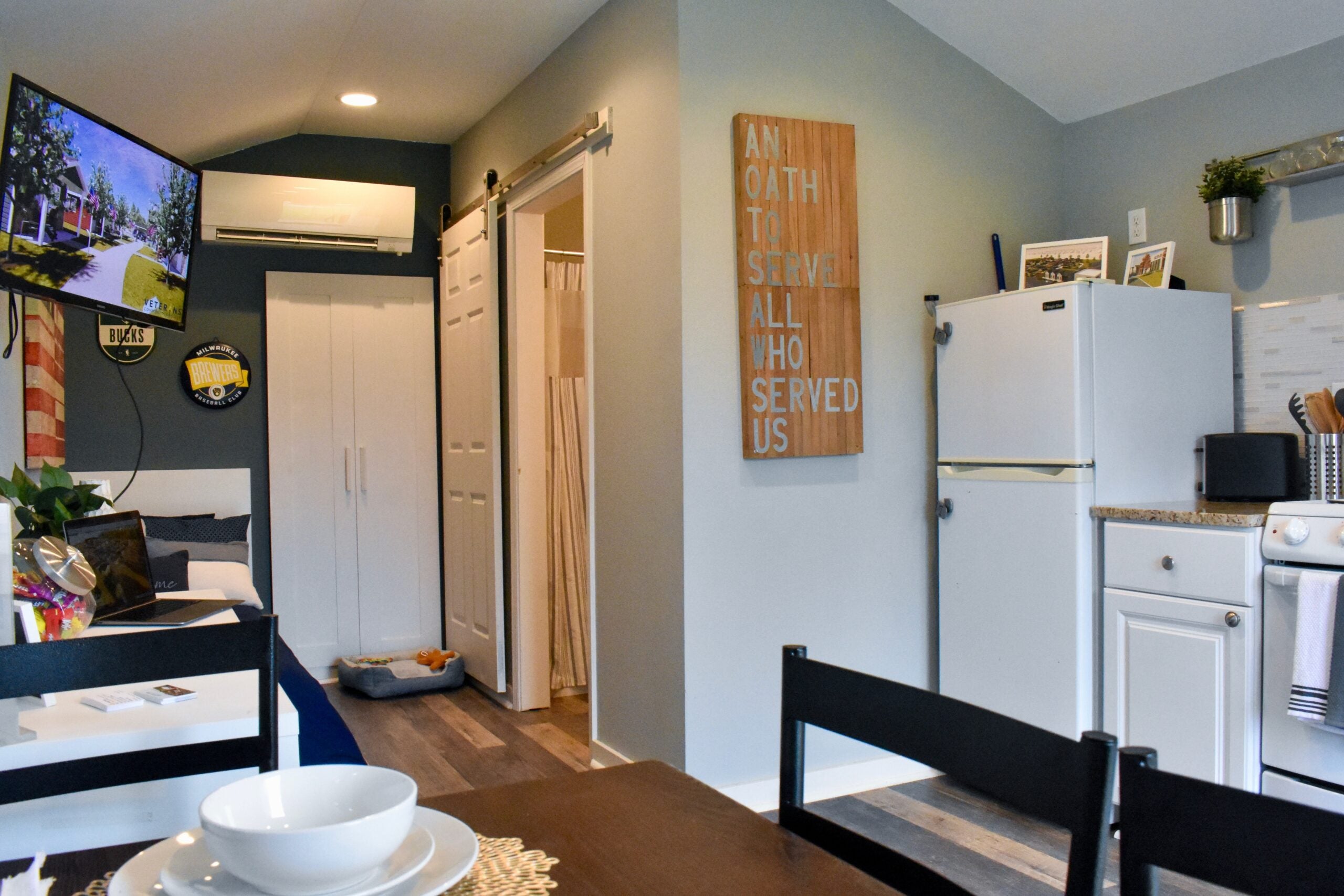 A small, modern studio apartment features a kitchenette, dining table, wall-mounted TV, and a sign reading “An oath to serve all who served us.”.