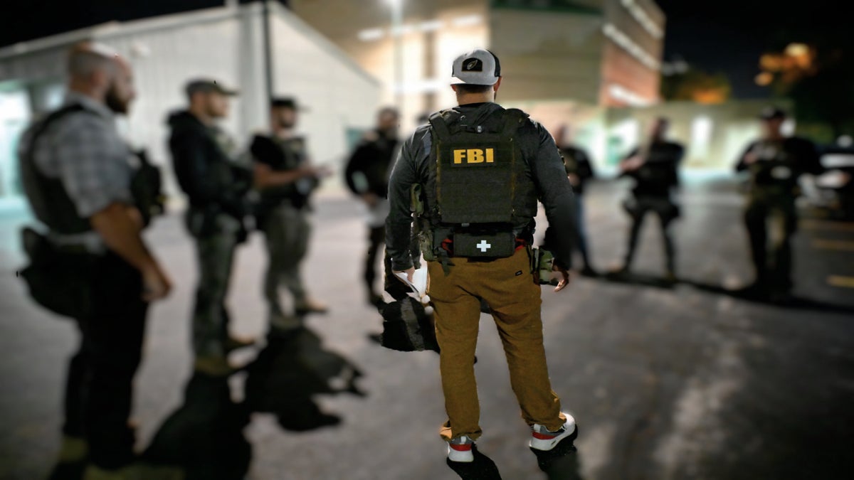 A group of armed FBI agents, one with FBI on his vest, stand together in a parking lot at night near a building.