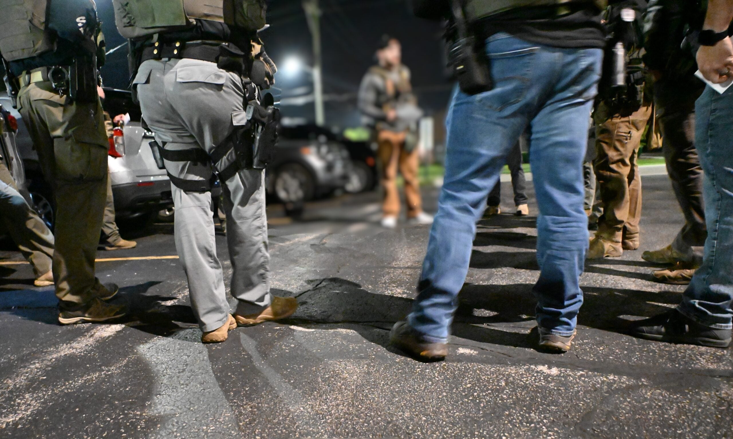 A group of law enforcement officers stand in a circle on a paved surface at night, with weapons and gear visible on their belts.