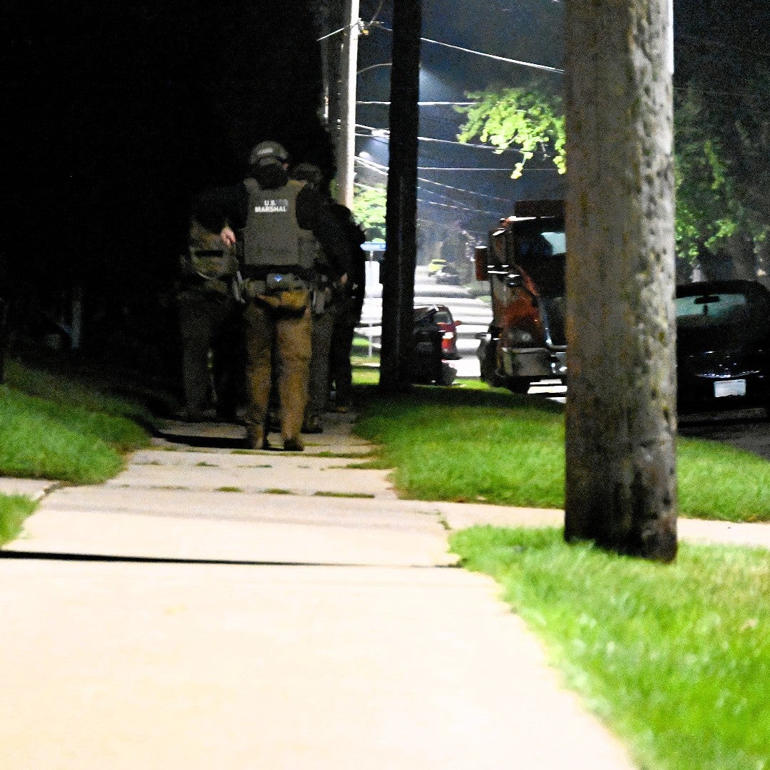 A group of armed law enforcement officers walk on a sidewalk at night, with a police vehicle and cars parked nearby.