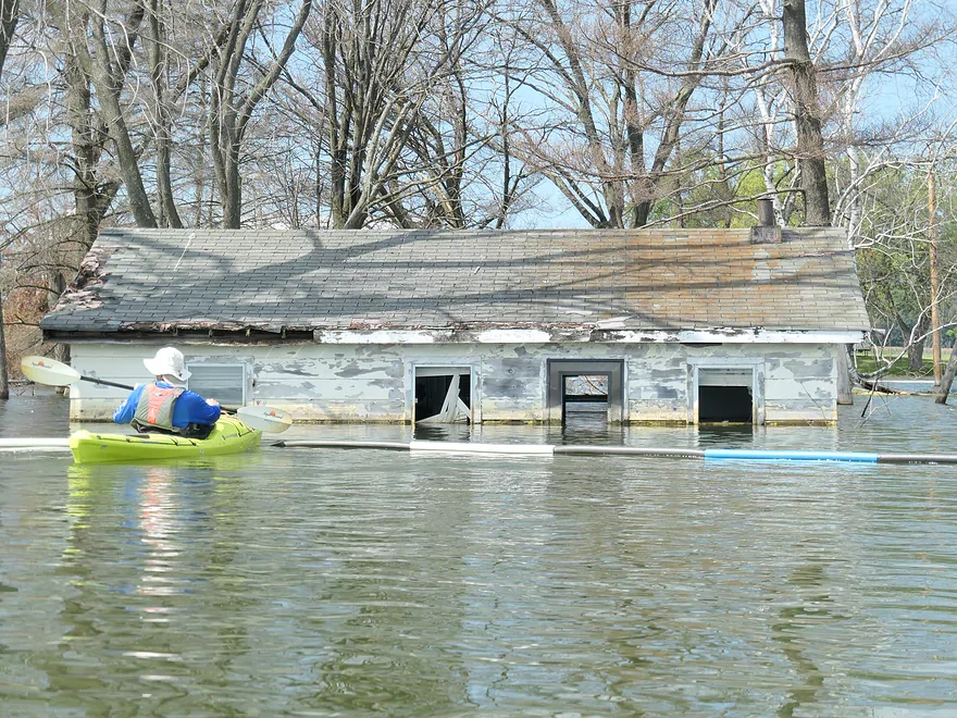 A person in a kayak paddles past a partially submerged, weathered house surrounded by floodwater and leafless trees.