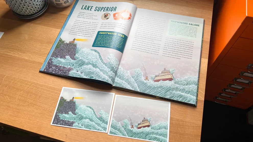 An open book about Lake Superior with illustrations of ships in stormy seas, accompanied by two printed cards with matching ship and lighthouse artwork on a wooden desk.
