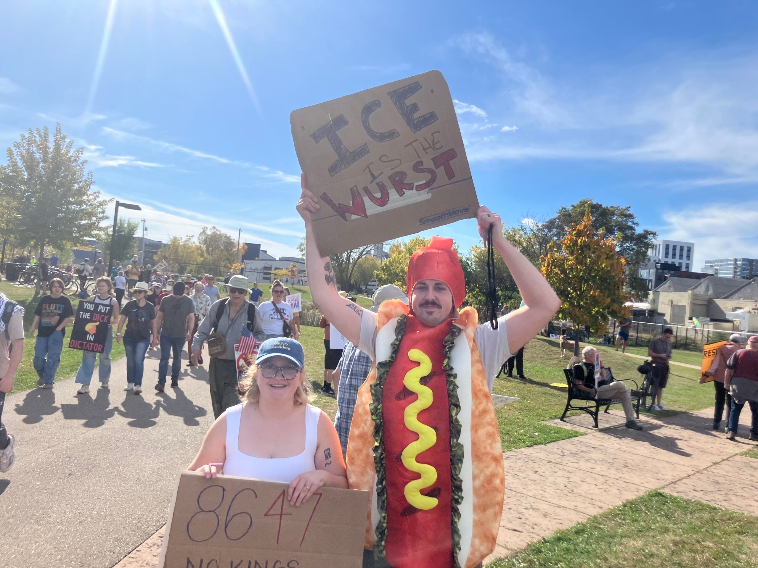 Two people at an outdoor event hold protest signs; one wears a hot dog costume and holds a sign that says, “ICE is the wurst.” Others are gathered in the background.