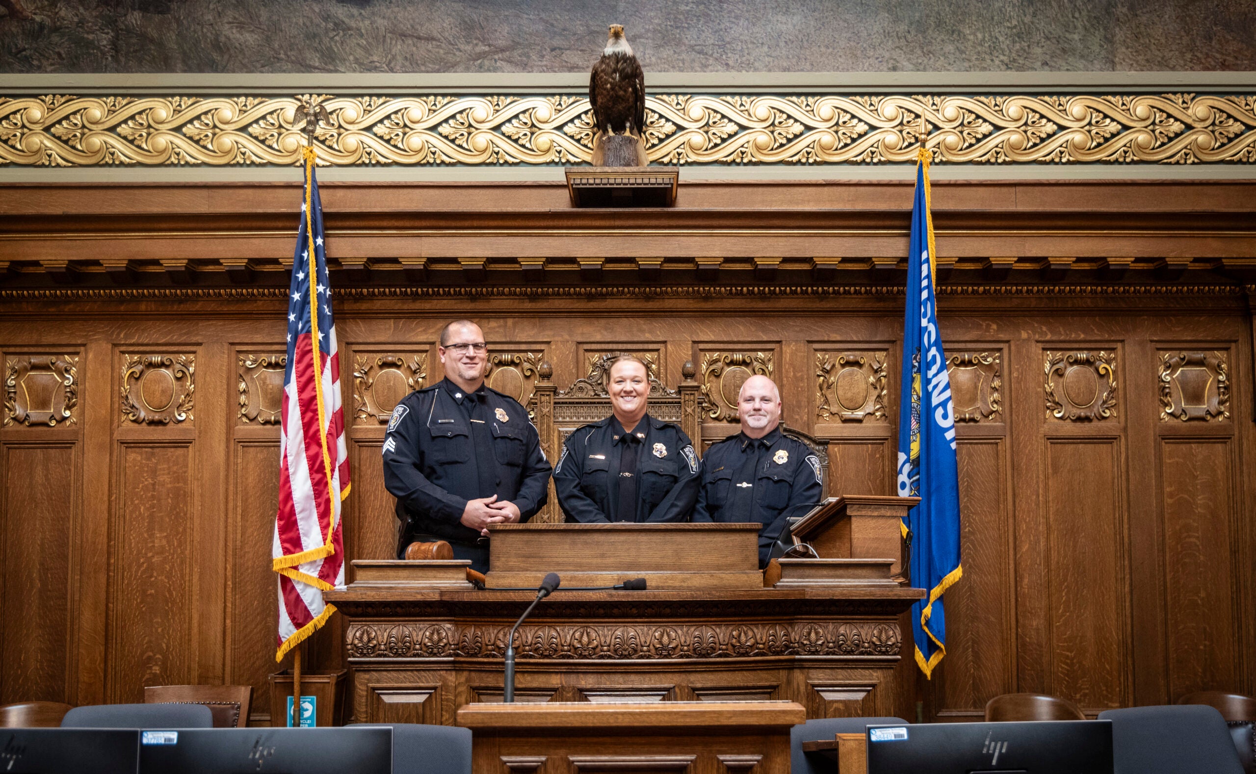 Three police officers stand side by side behind a wooden podium in a formal chamber, flanked by U.S. and state flags, with an eagle sculpture above them.