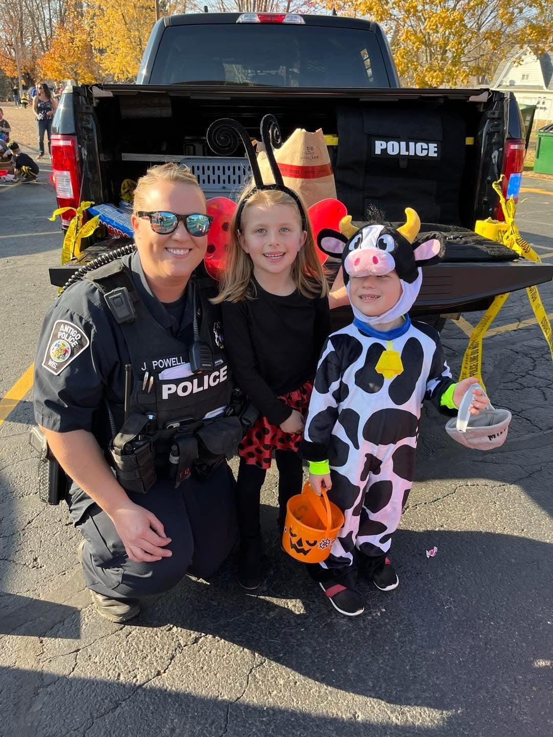 A police officer kneels next to two children in Halloween costumes, with a police vehicle and Halloween decorations in the background.