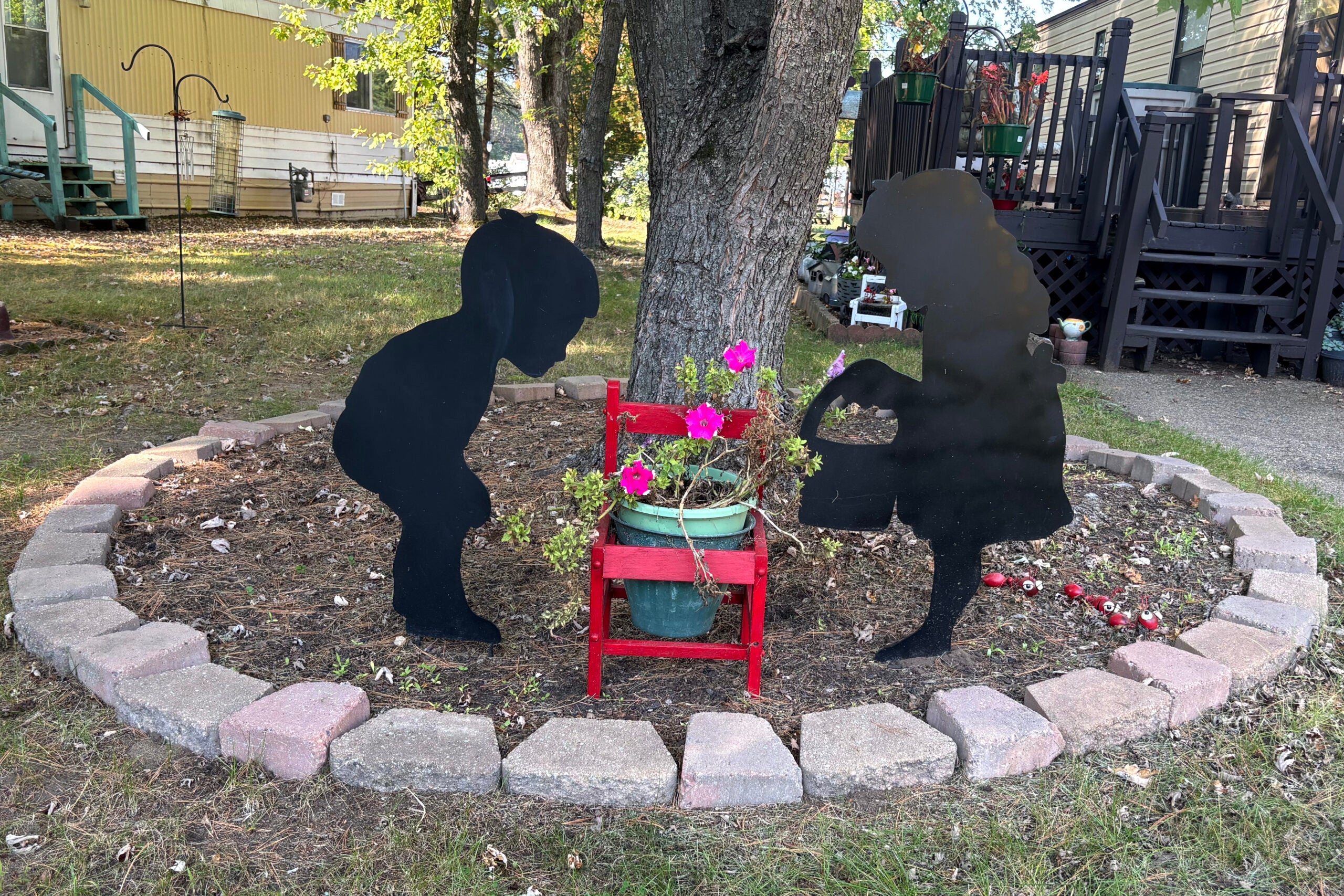 Two black silhouette cutouts of children face each other in a garden bed with a red chair holding a potted plant with pink flowers, surrounded by a brick border.