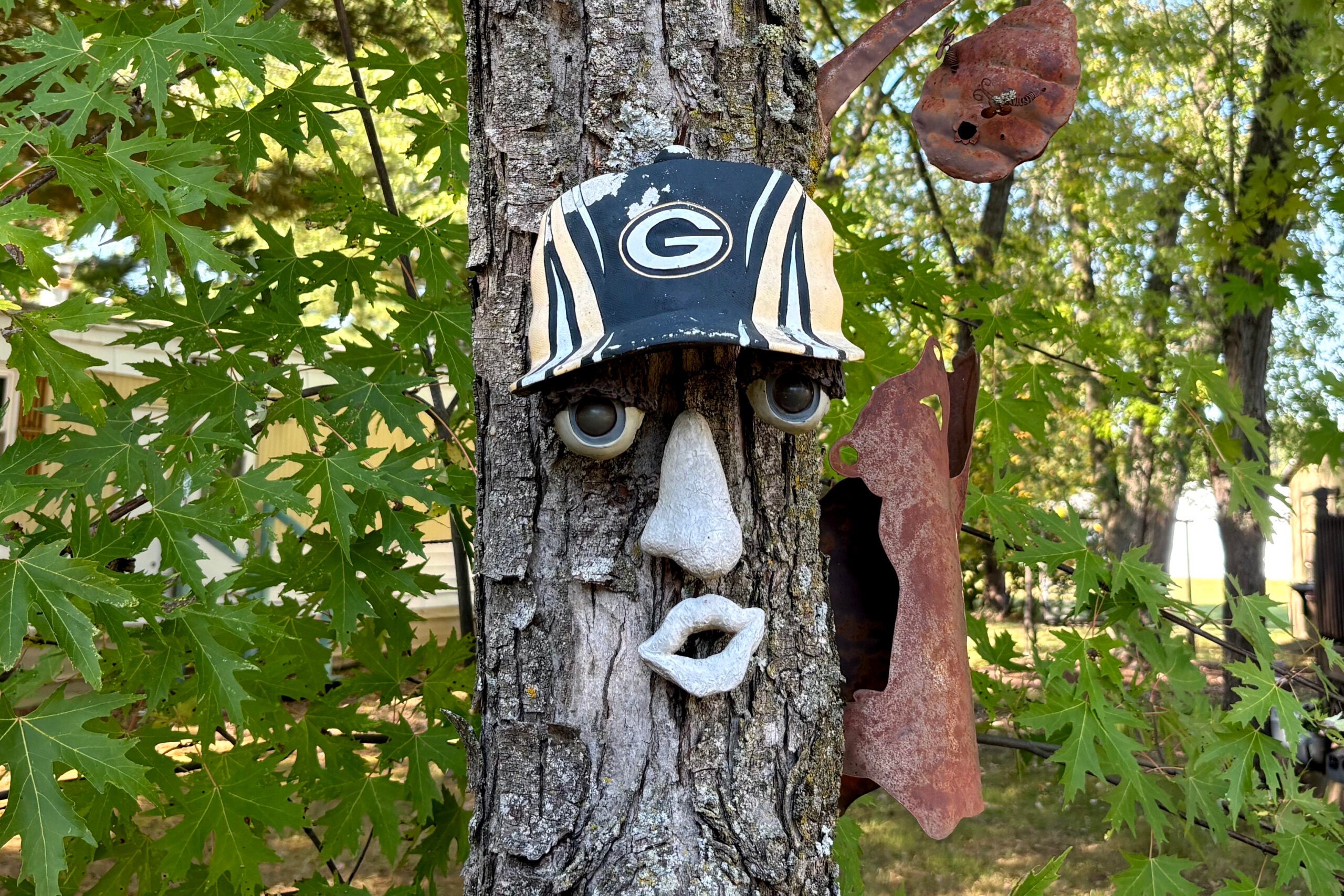 A tree trunk decorated with a face and a Green Bay Packers cap, with eyes, nose, and mouth sculptures attached, in a leafy outdoor setting.
