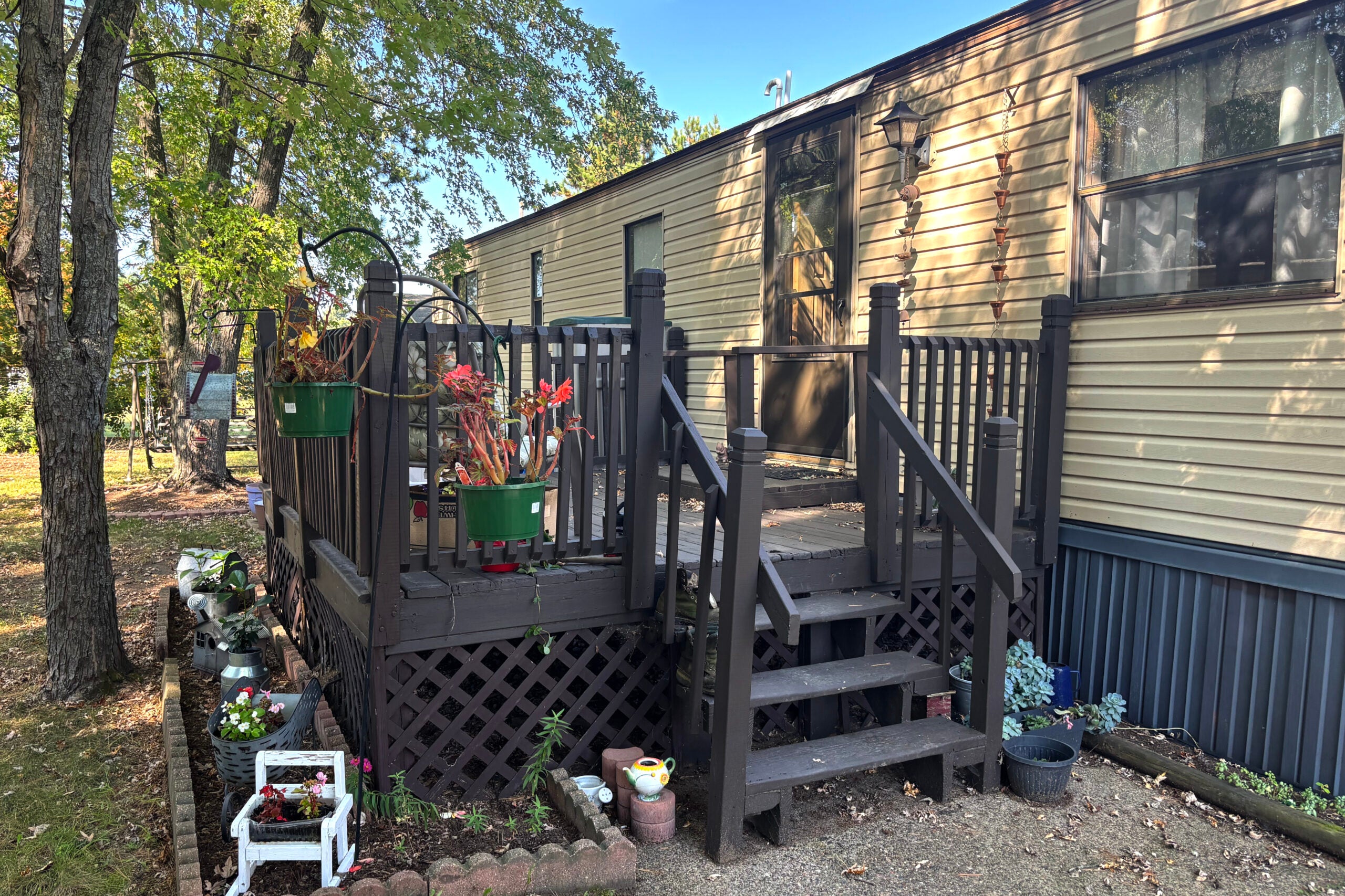 A wooden porch with steps and railing is attached to a beige mobile home, surrounded by potted plants, flower boxes, and trees.