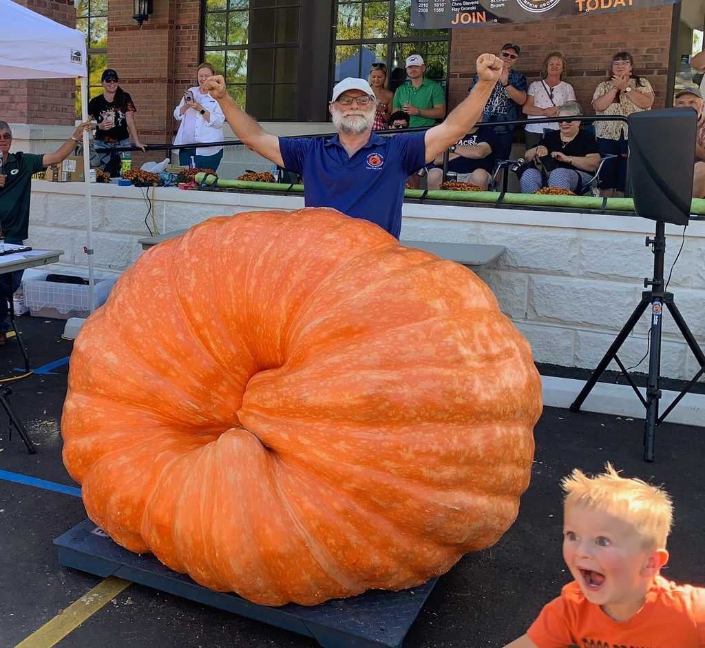 A man stands with raised arms behind an enormous pumpkin at an outdoor event, while a young child reacts excitedly in the foreground.