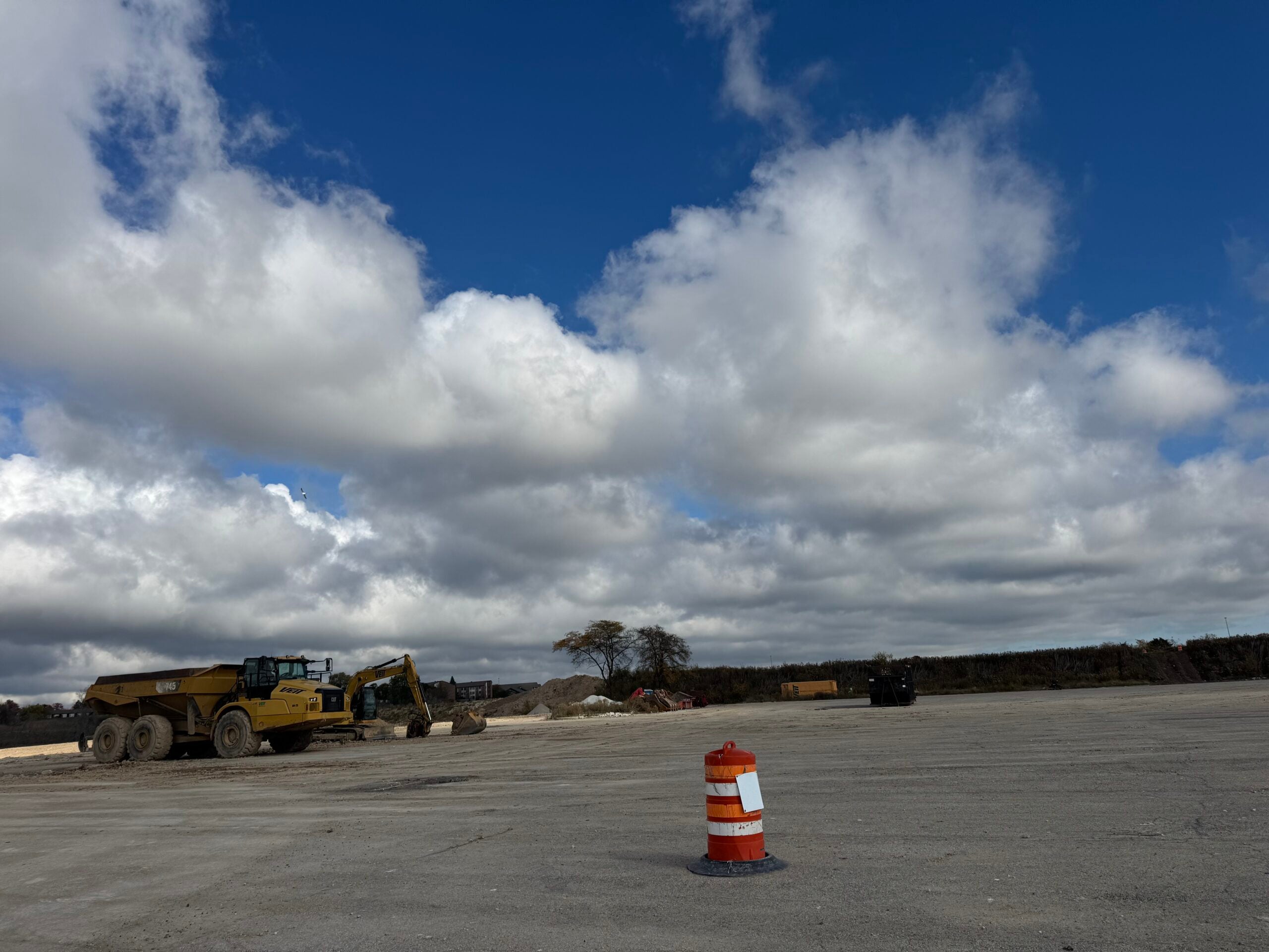 An orange and white traffic barrel stands on an empty lot with construction vehicles and equipment in the background under a partly cloudy sky.