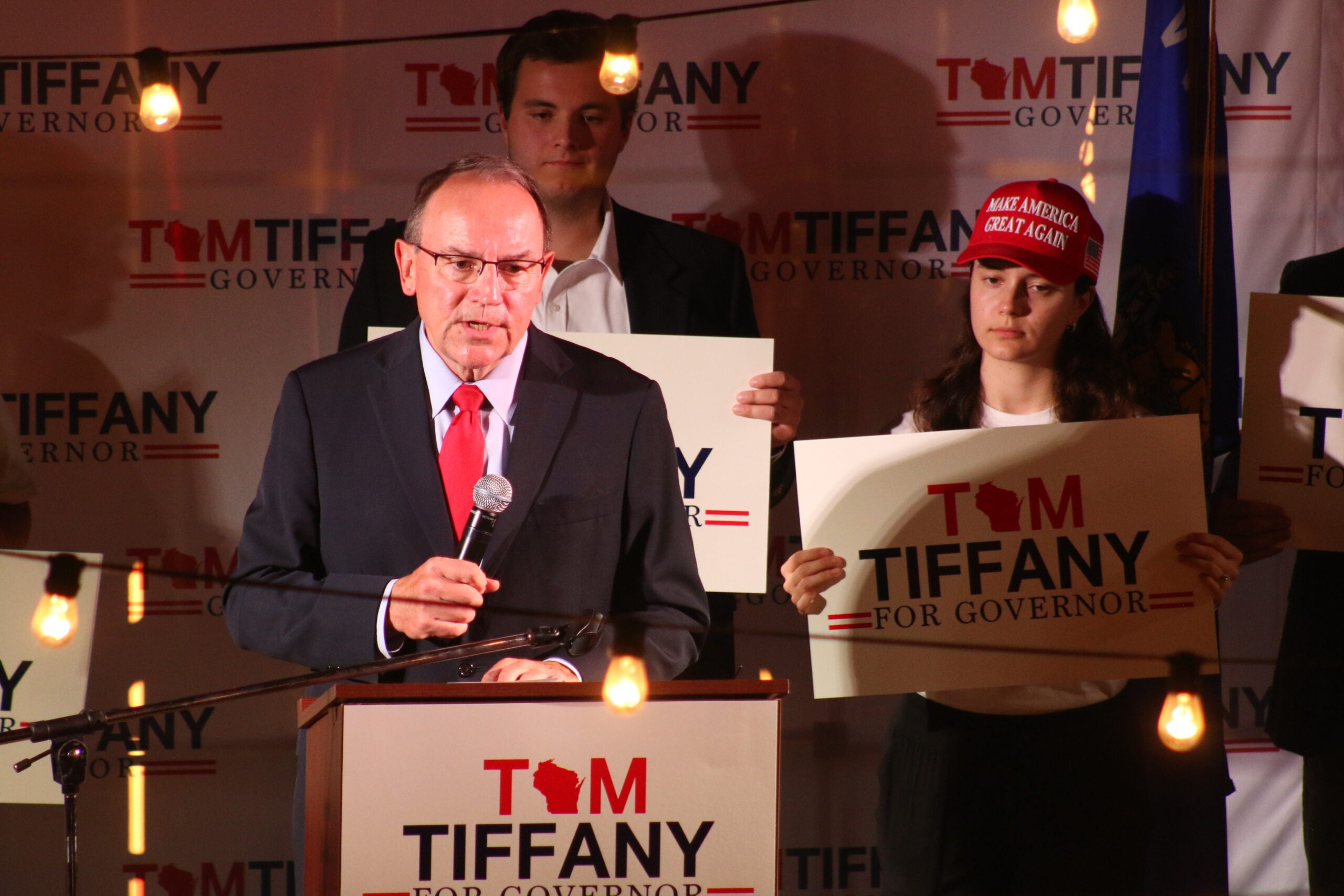 A man in a suit speaks at a podium with Tiffany for Governor signs behind him; supporters stand nearby, one wearing a Make America Great Again hat.