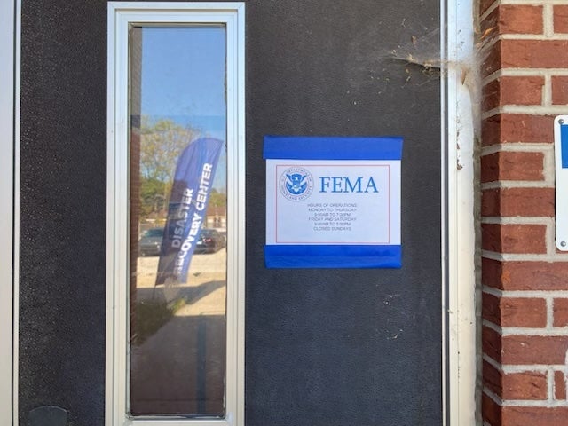 A door with a sign taped to it labeled FEMA and information about hours and operations. A Disaster Recovery Center flag is visible through the window.