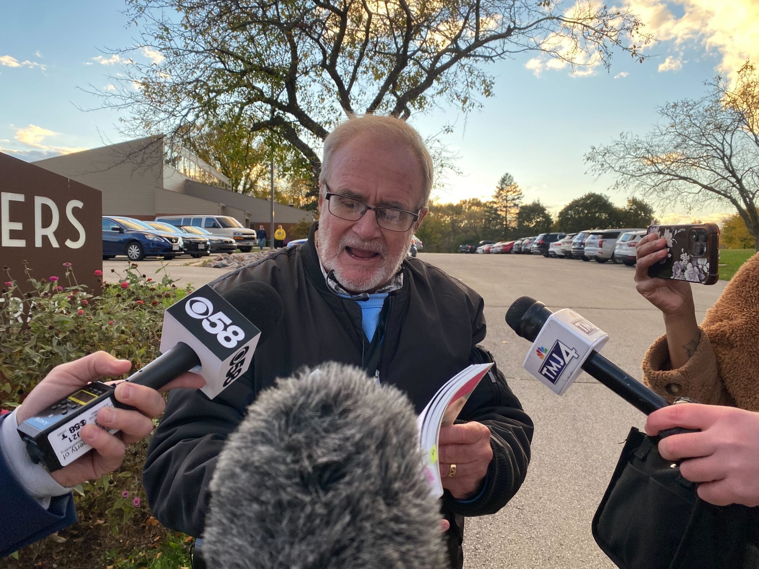 An older man speaks to reporters outside, holding a booklet while multiple microphones are pointed toward him. Cars are parked in the background.