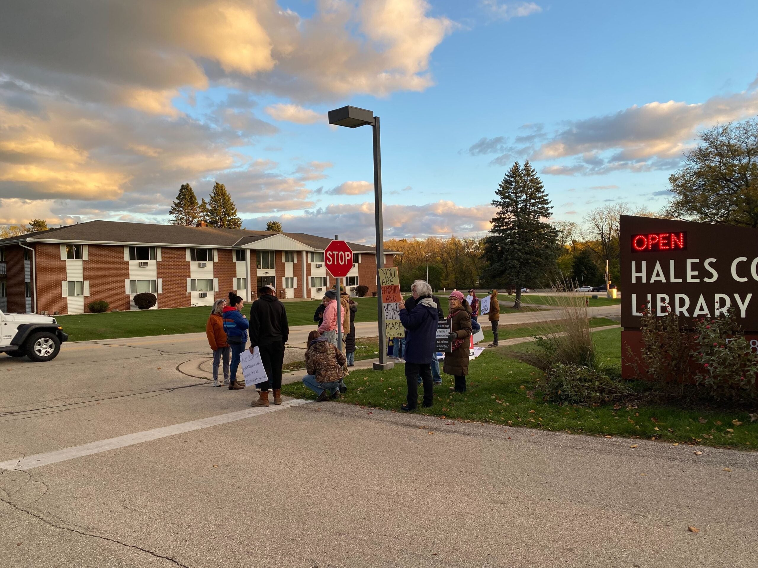 A group of people stands near a stop sign and the entrance to Hales Corners Library as cars pass by on a residential street at sunset.