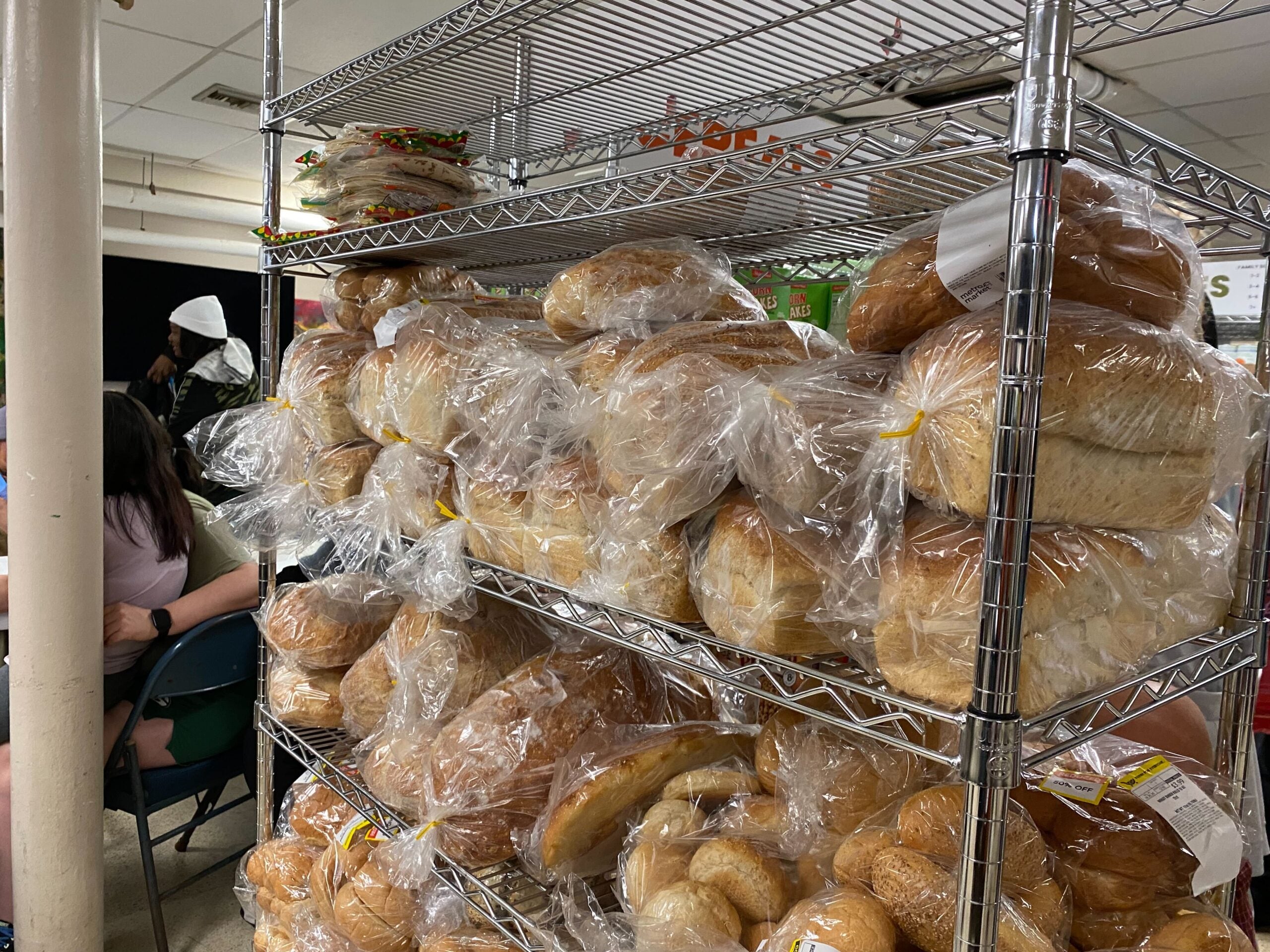 Metal shelves stacked with loaves of bread and rolls wrapped in plastic; people are seated and visible in the background.