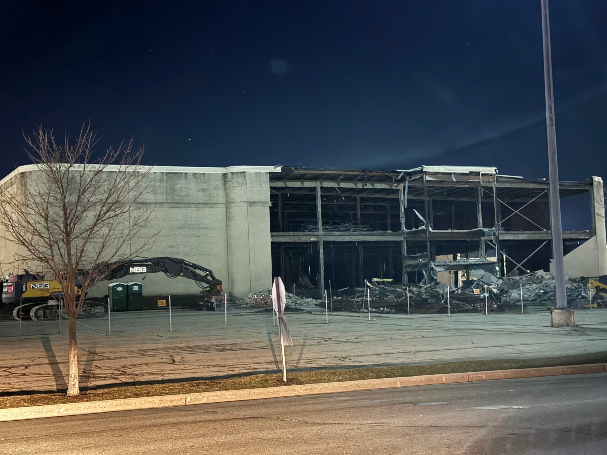 A partially demolished building at night, with debris visible inside and a construction excavator parked outside. The area is surrounded by a temporary fence.