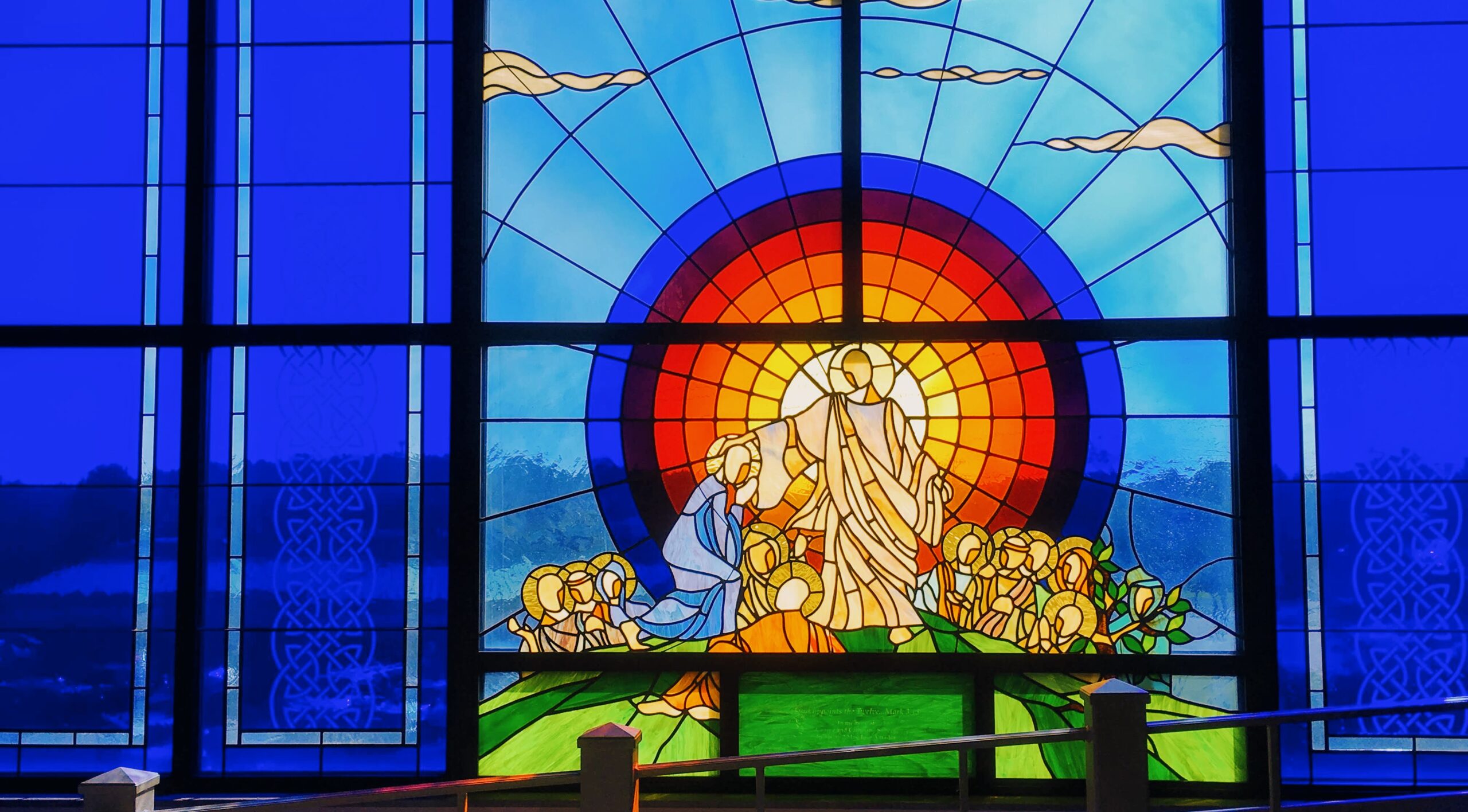 Stained glass window depicting Jesus teaching a group of people, with a radiant sun and blue sky in the background.