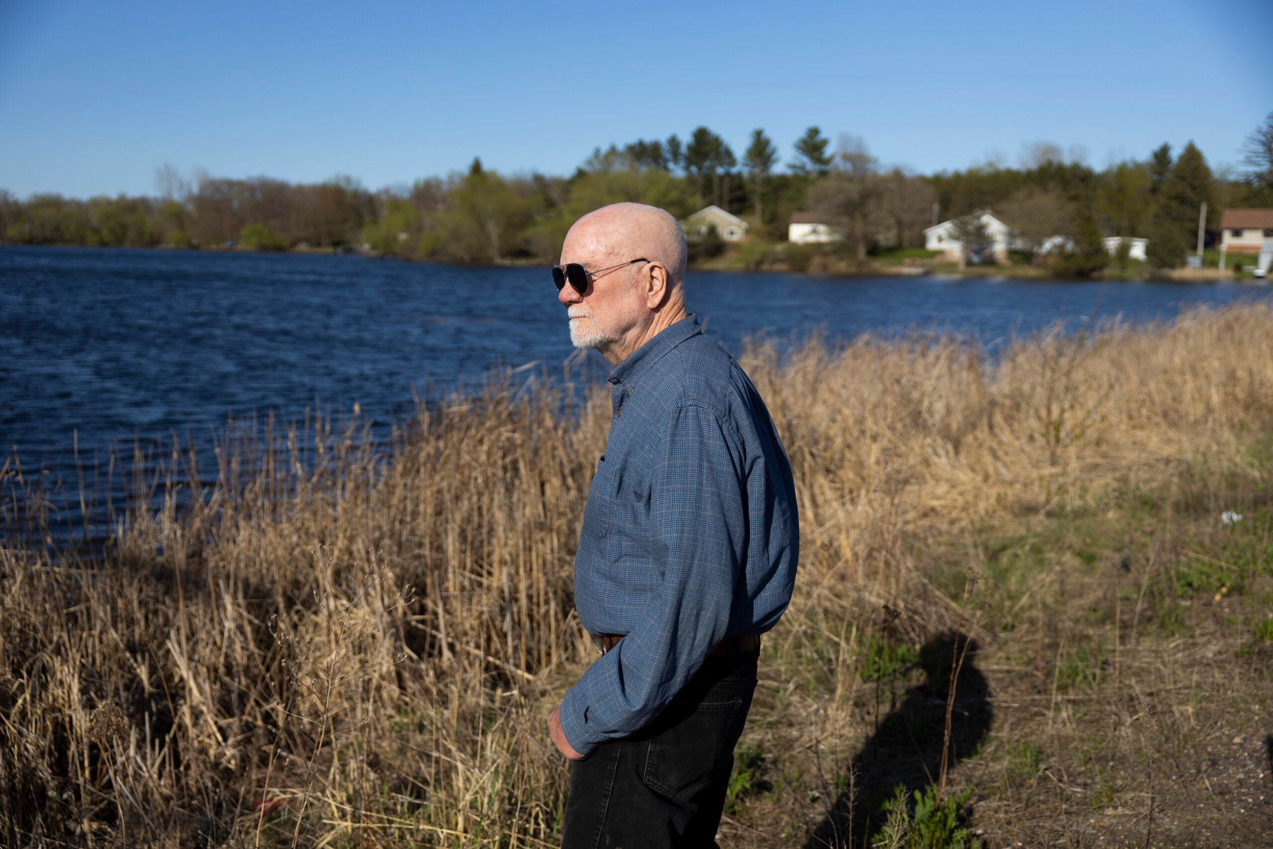 An older man wearing sunglasses and a blue shirt stands by a lake, surrounded by dry grass and trees, with houses visible in the background under a clear sky.
