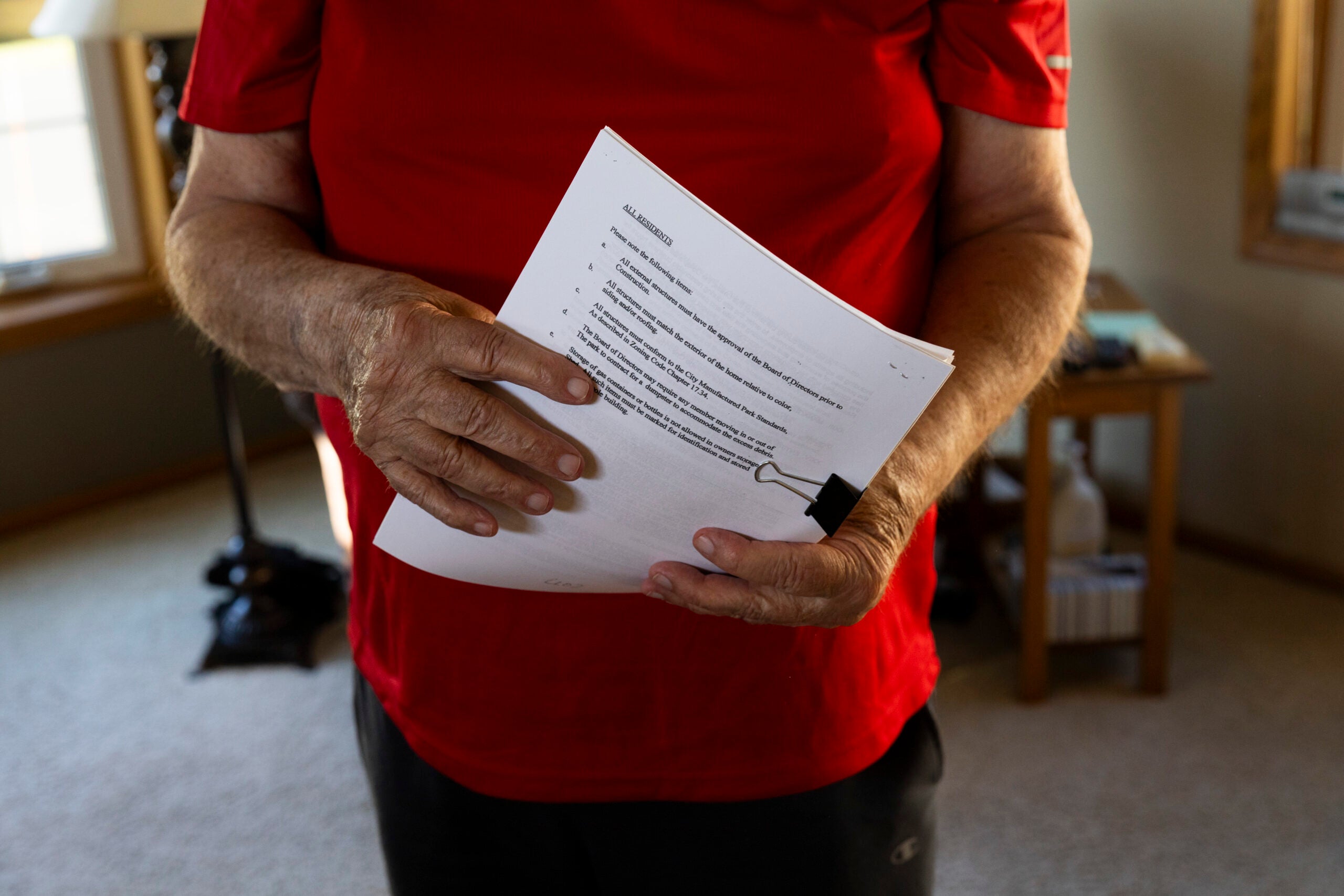 A person in a red shirt holds a printed document stapled with a binder clip, standing in a sunlit room with furniture in the background.