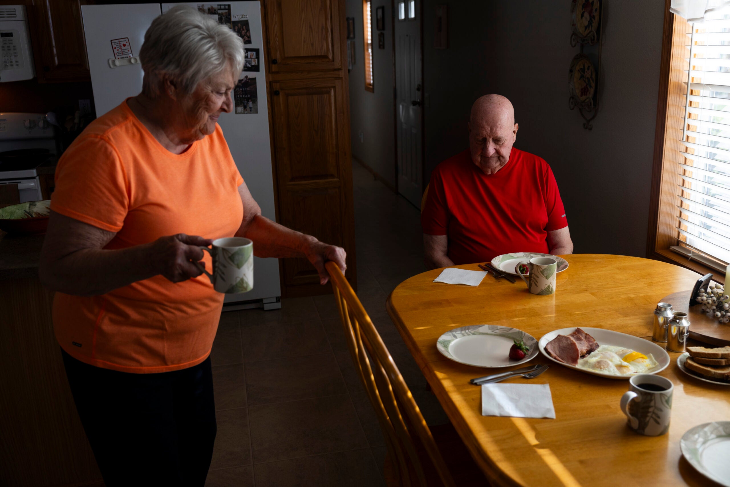 Two older adults sit at a wooden dining table with plates of food and coffee mugs in a warmly lit kitchen.