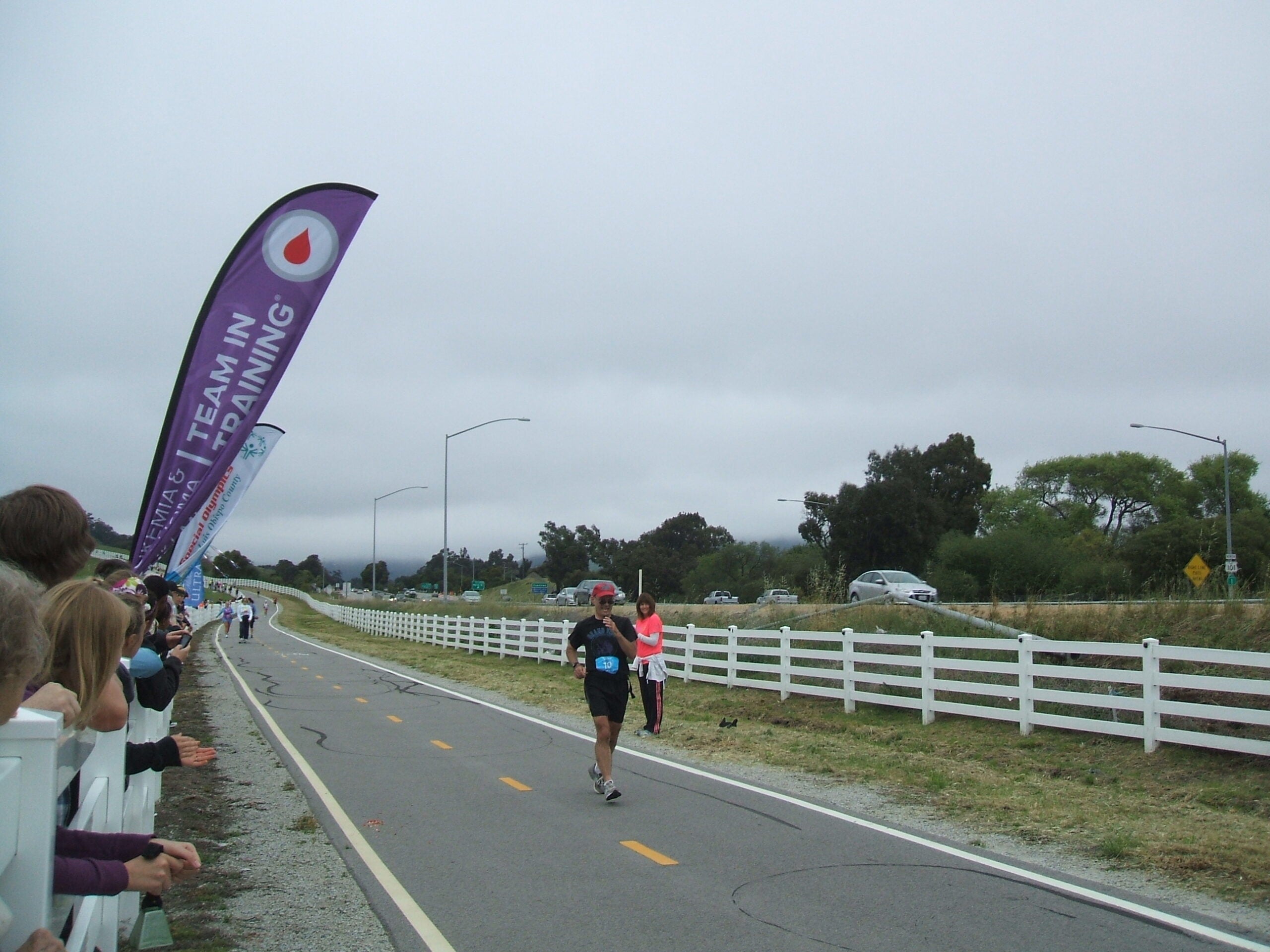 A runner approaches the finish line on a paved path while spectators cheer, with a Team in Training banner and overcast sky in the background.