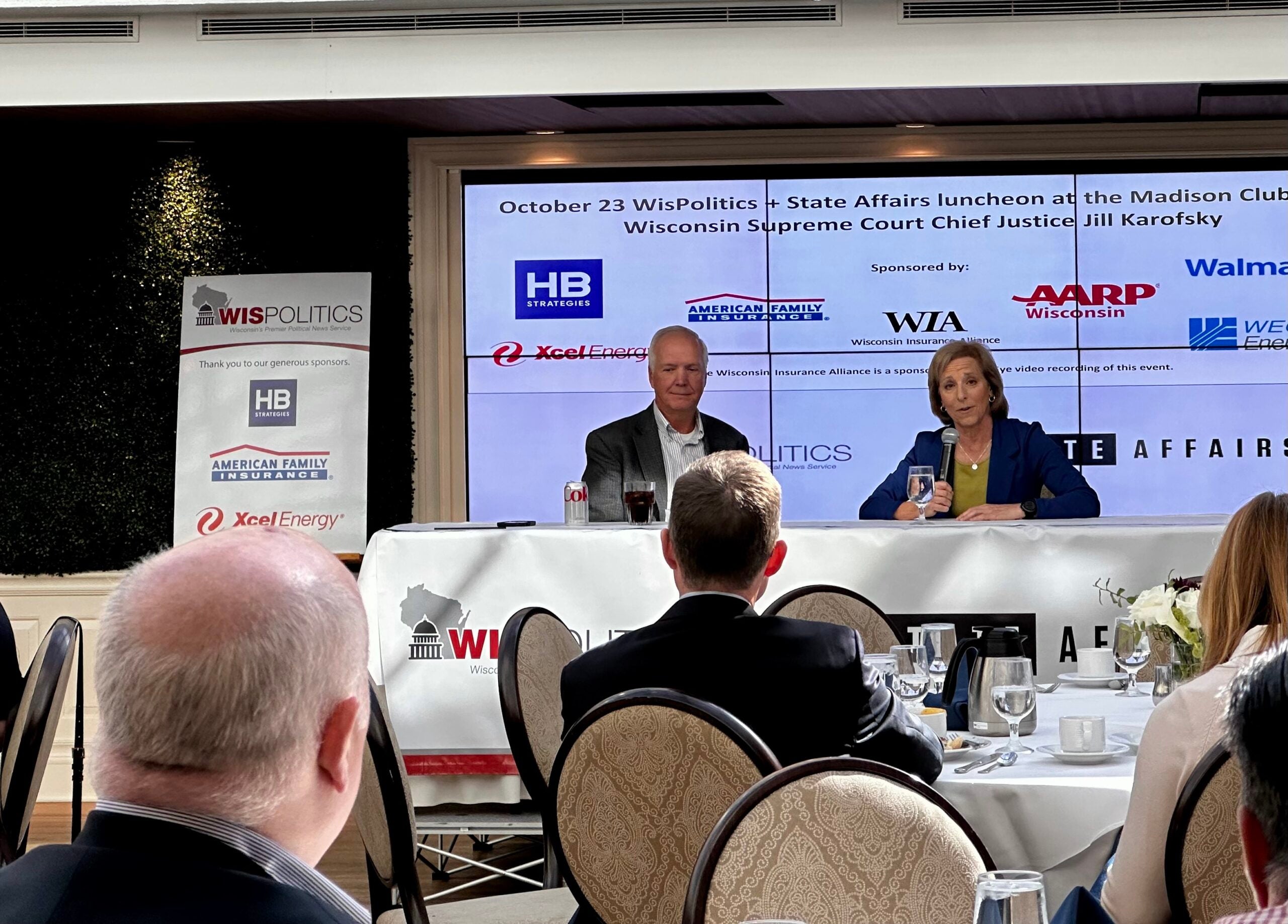 Two speakers sit at a table on stage at a WisPolitics State Affairs luncheon, with event banners and a presentation screen in the background.