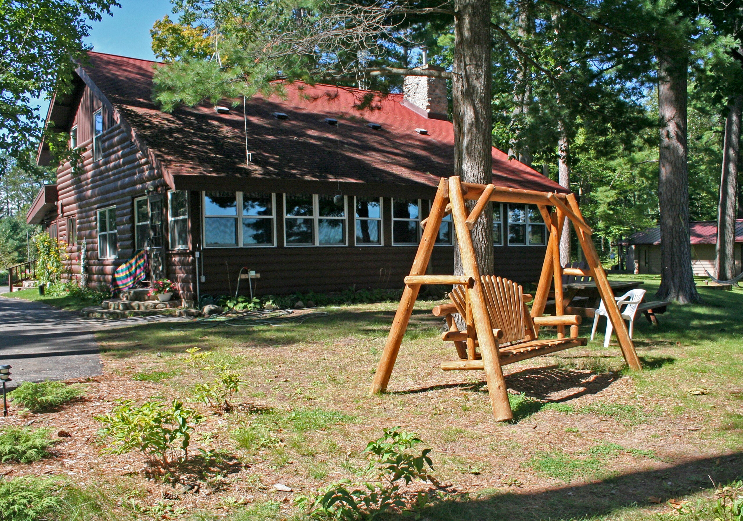 A wooden swing set stands in the yard of a brown log cabin surrounded by trees, with lawn chairs and plants visible nearby.