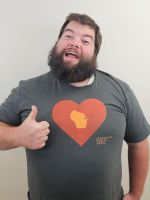 A man with a beard smiles and gives a thumbs up while wearing a gray t-shirt with a large red heart and a yellow shape in the center.