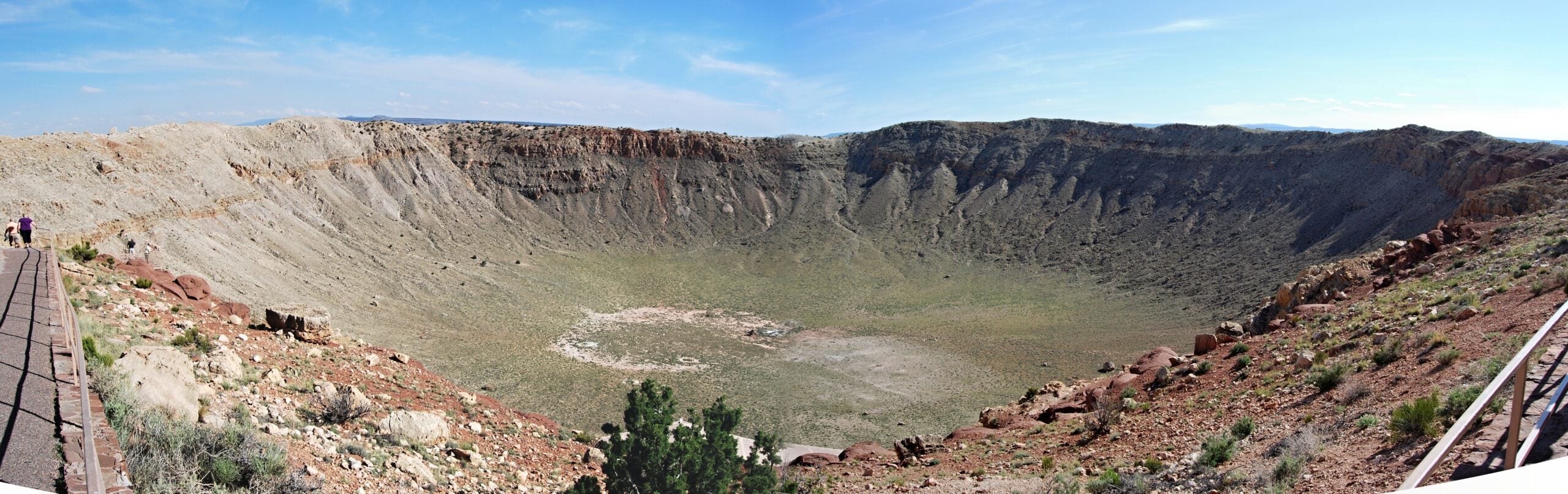 Panoramic view of a large meteor crater with rocky walls and sparse vegetation, bordered by railings and a walkway on the left side.