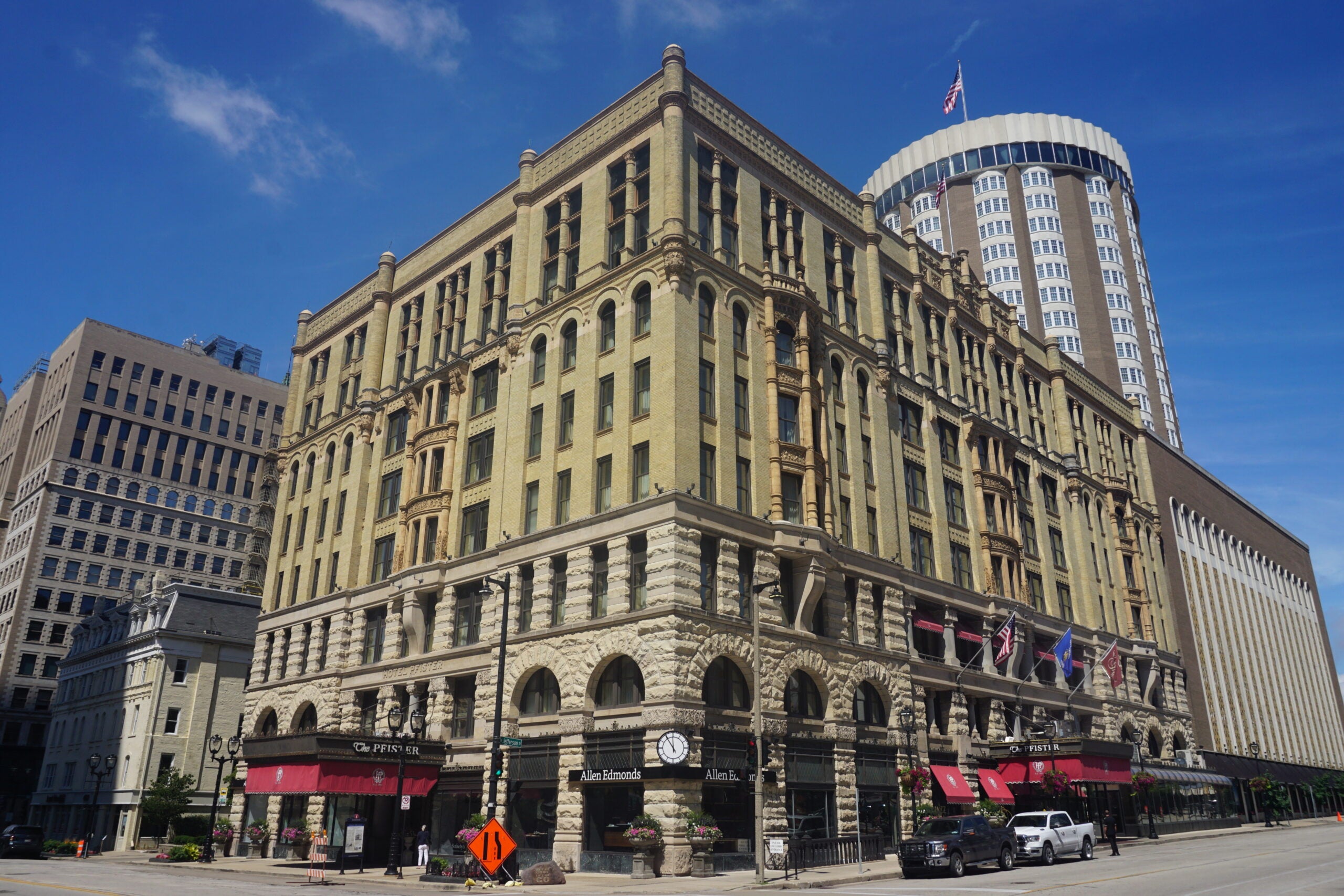 A historic multi-story stone hotel with arched windows and several flags at the entrance, located on a city street with modern buildings in the background.