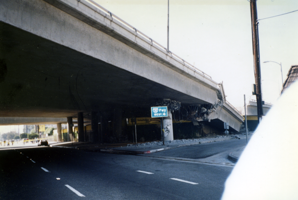 A damaged freeway overpass with a collapsed section above a road, debris scattered below, and a green highway sign for I-5 visible.