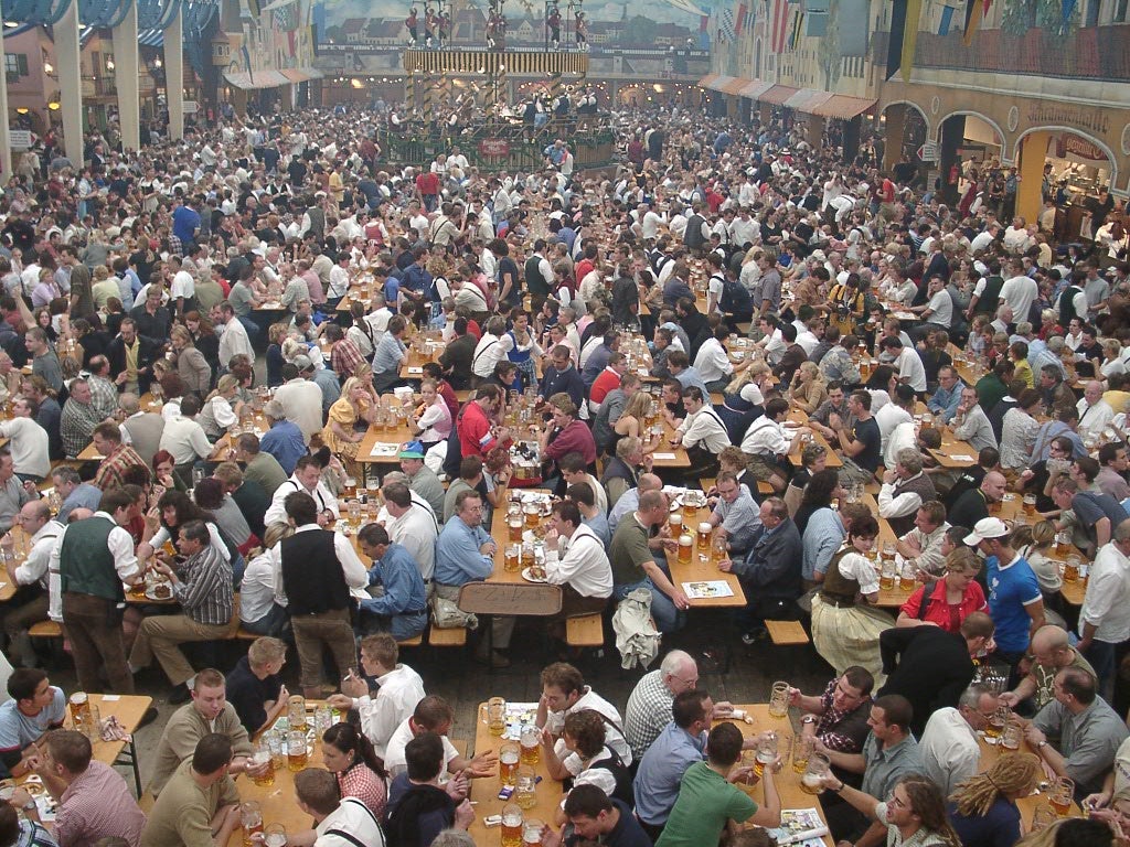 Large crowd of people sit at long wooden tables inside a hall, drinking beer and socializing at a festival event.