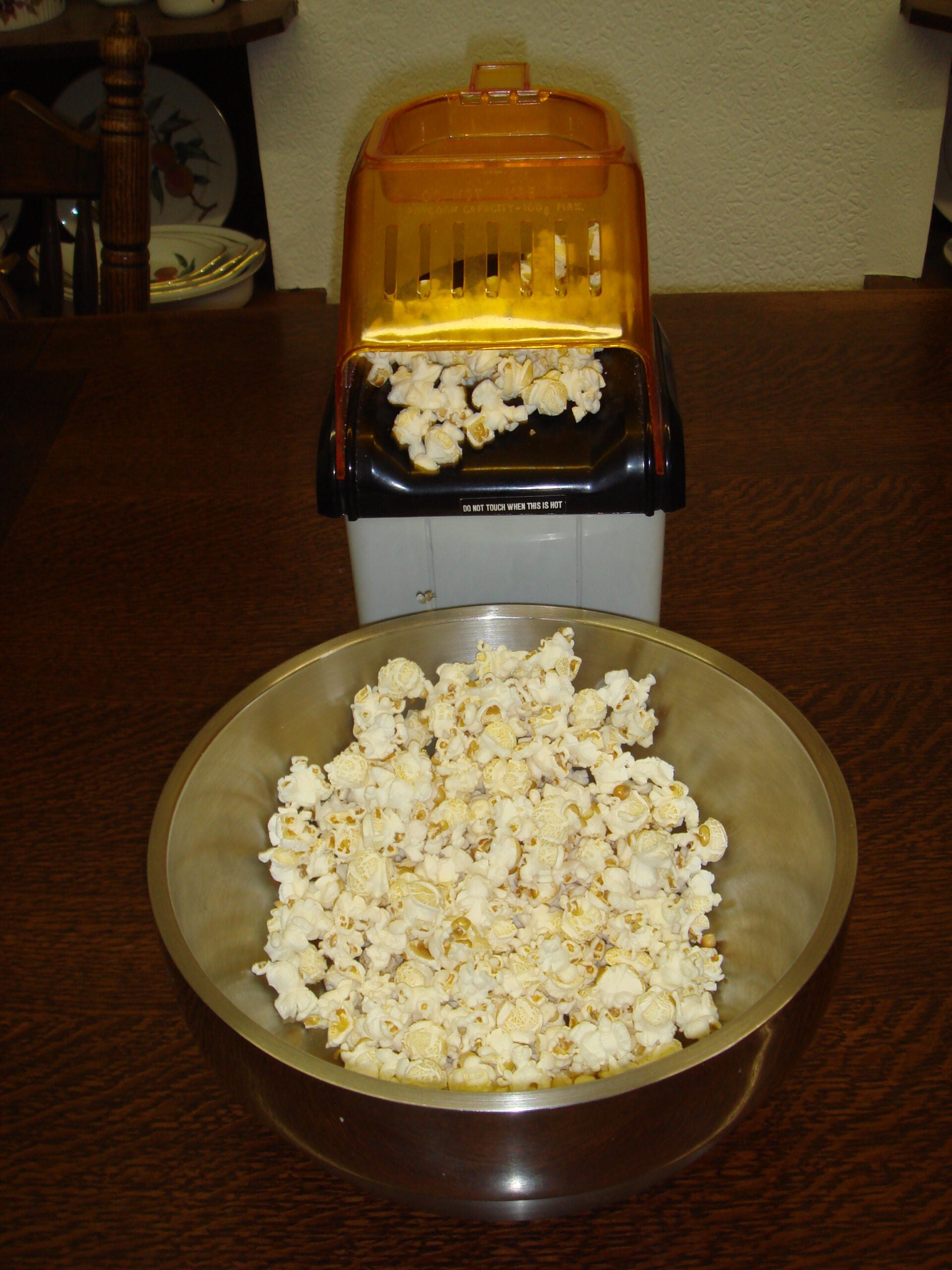 A metal bowl filled with popcorn sits in front of a popcorn maker on a wooden table.