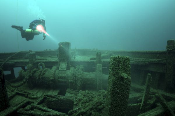 A scuba diver with a flashlight explores the moss-covered remains of a sunken shipwreck underwater.