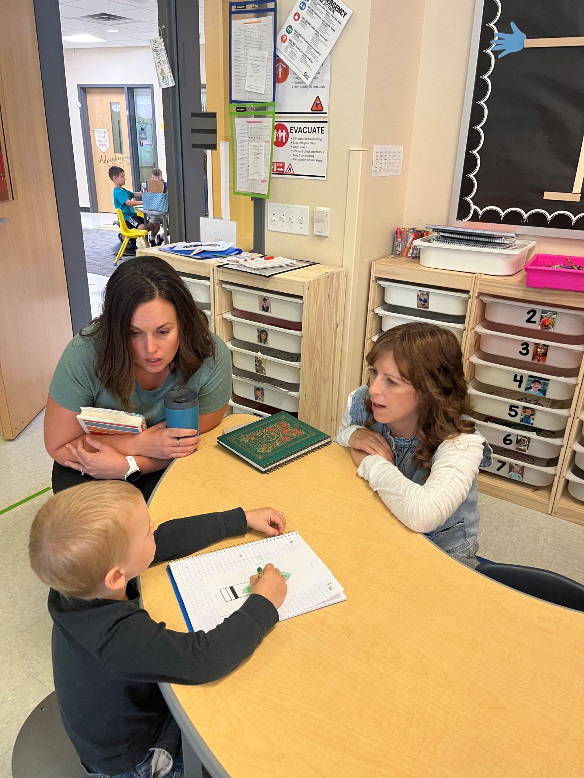 A woman and a girl sit at a table with a young boy who is writing in a notebook in a classroom. Shelves with bins and educational posters are visible in the background.