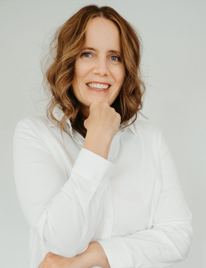 A woman with wavy brown hair wearing a white button-up shirt smiles at the camera with her hand resting under her chin against a plain background.