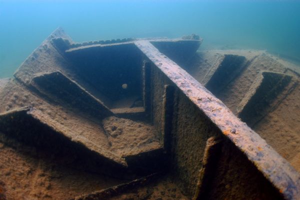 Large, rusted metal paddle wheel submerged underwater, covered in algae and sediment, with soft natural light filtering through.