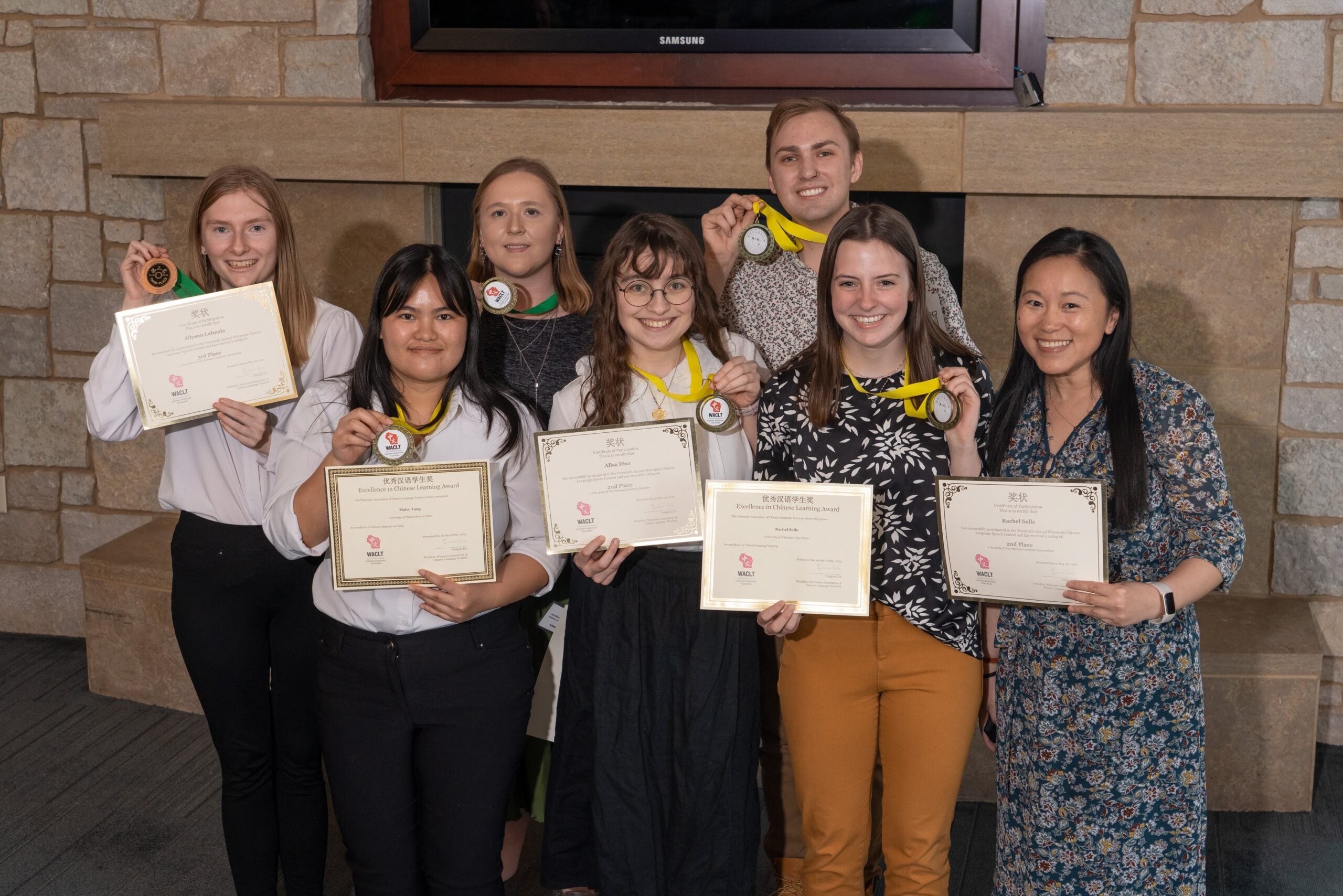 Seven people pose indoors holding certificates and medals, standing in front of a stone wall and fireplace, smiling at the camera.