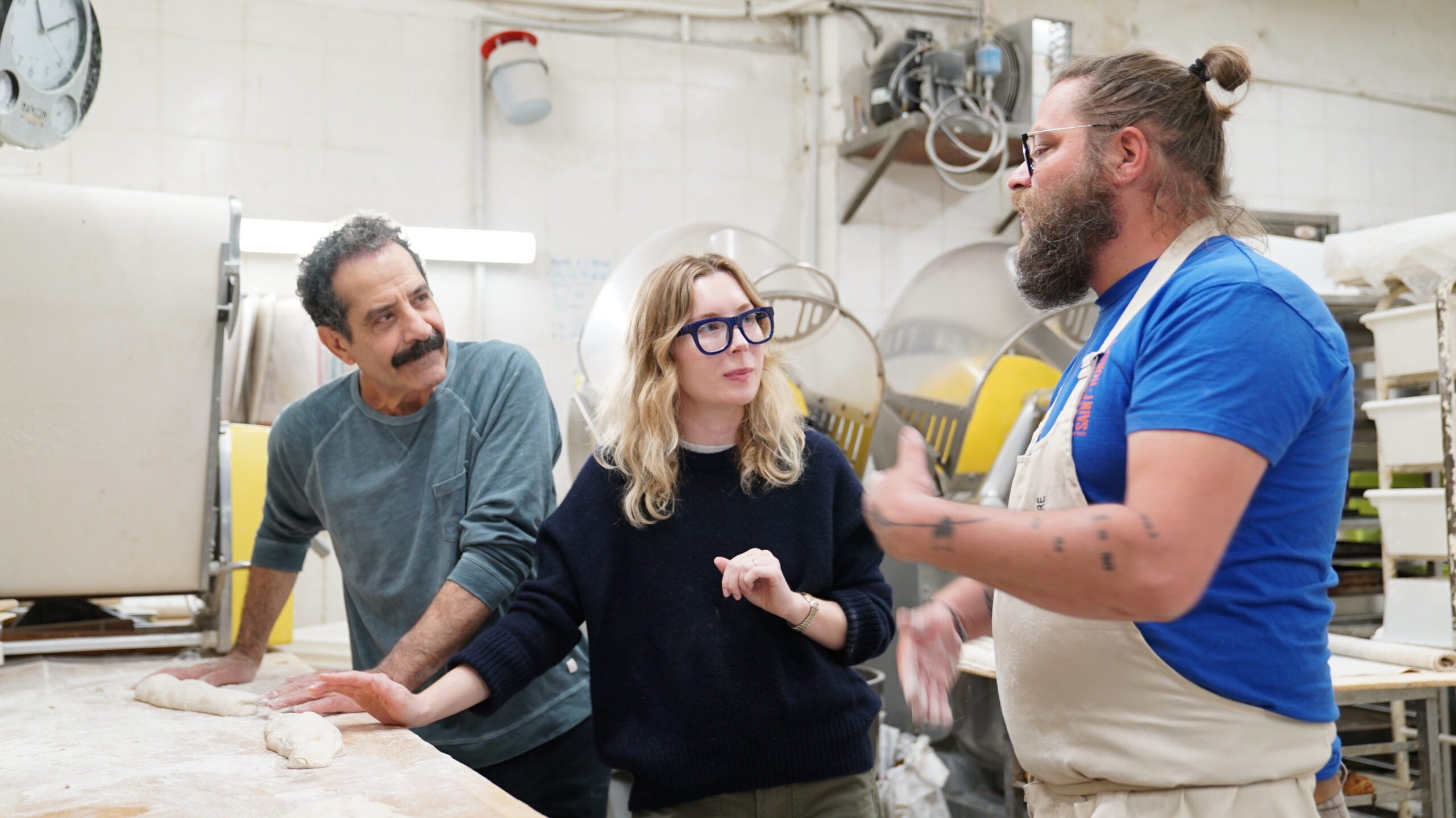 Three people stand in a bakery kitchen; one explains something while the others listen, with dough and baking equipment visible on the counter.