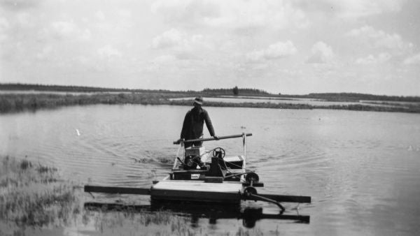 A person stands on a small, flat, mechanical boat or raft with equipment, navigating through a calm, shallow body of water with grassy banks.