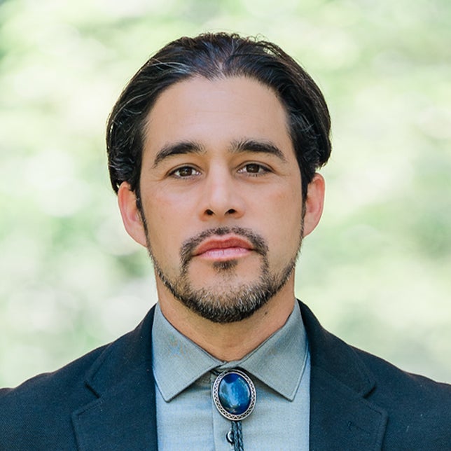 A man with dark hair and a goatee wearing a dark jacket, light shirt, and a blue bolo tie stands in front of a blurred outdoor background.