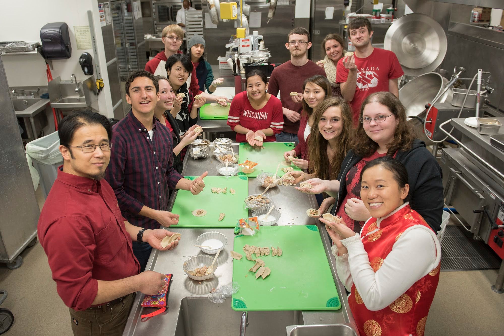A group of people stands around a kitchen island in a commercial kitchen, smiling and making dumplings together.