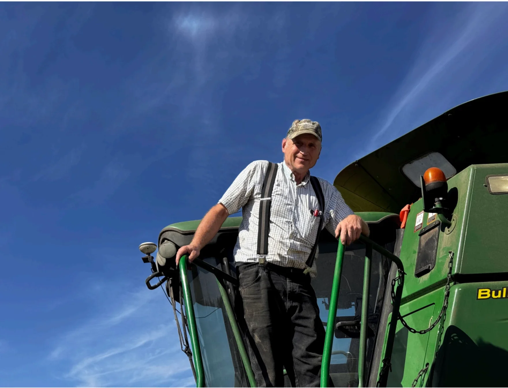 An older man wearing a cap and suspenders stands on the steps of a large green agricultural machine under a bright blue sky.