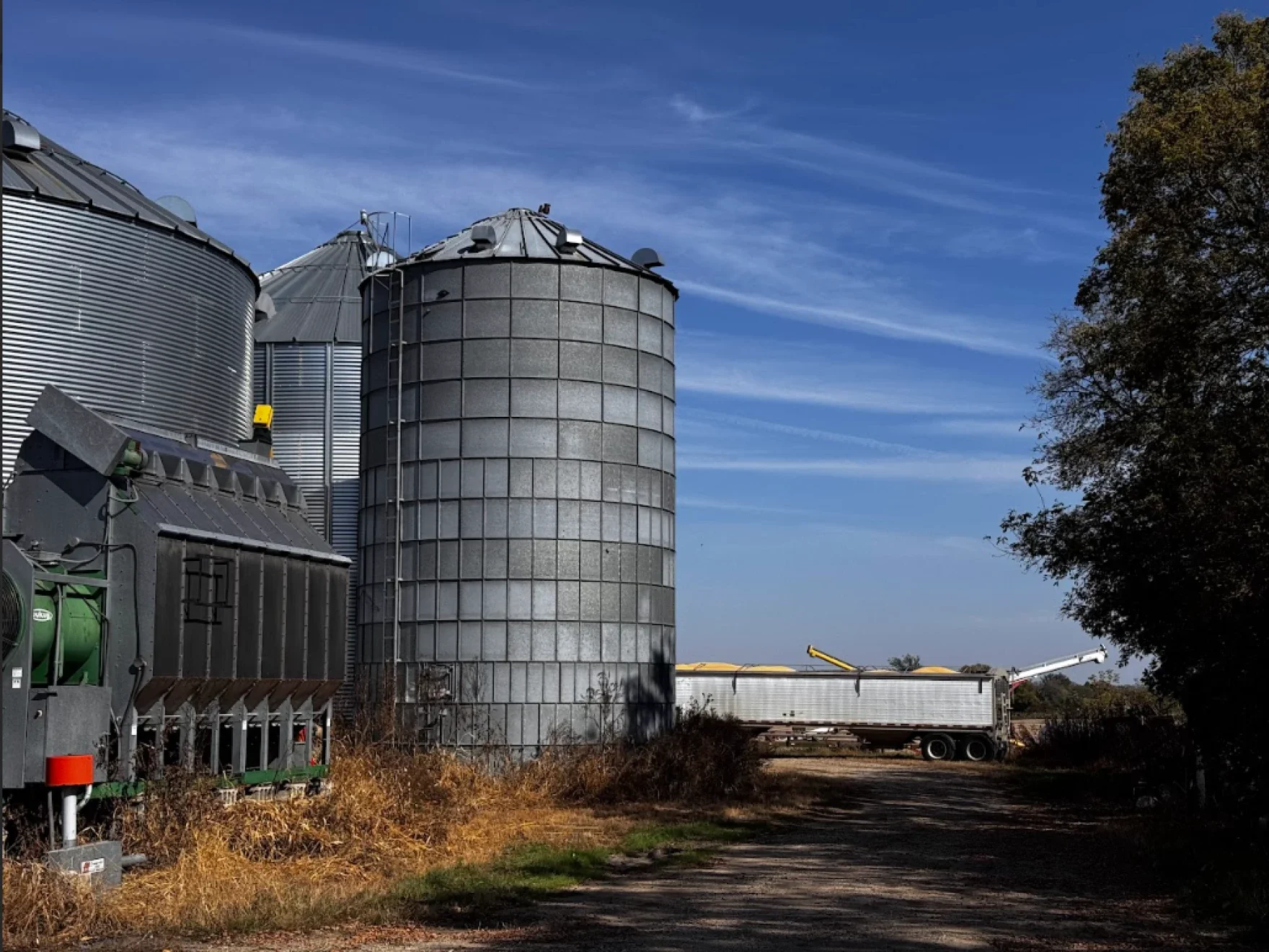 Large metal grain silos stand beside a farm road with a white semi-trailer truck parked nearby under a blue sky.