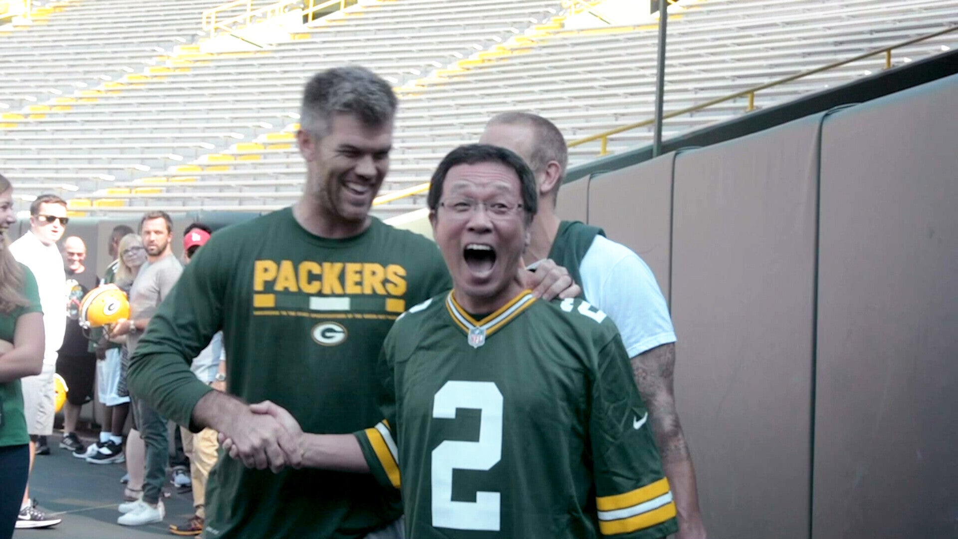 Two men in Green Bay Packers jerseys smile and shake hands on a football field, with empty stadium seats in the background.