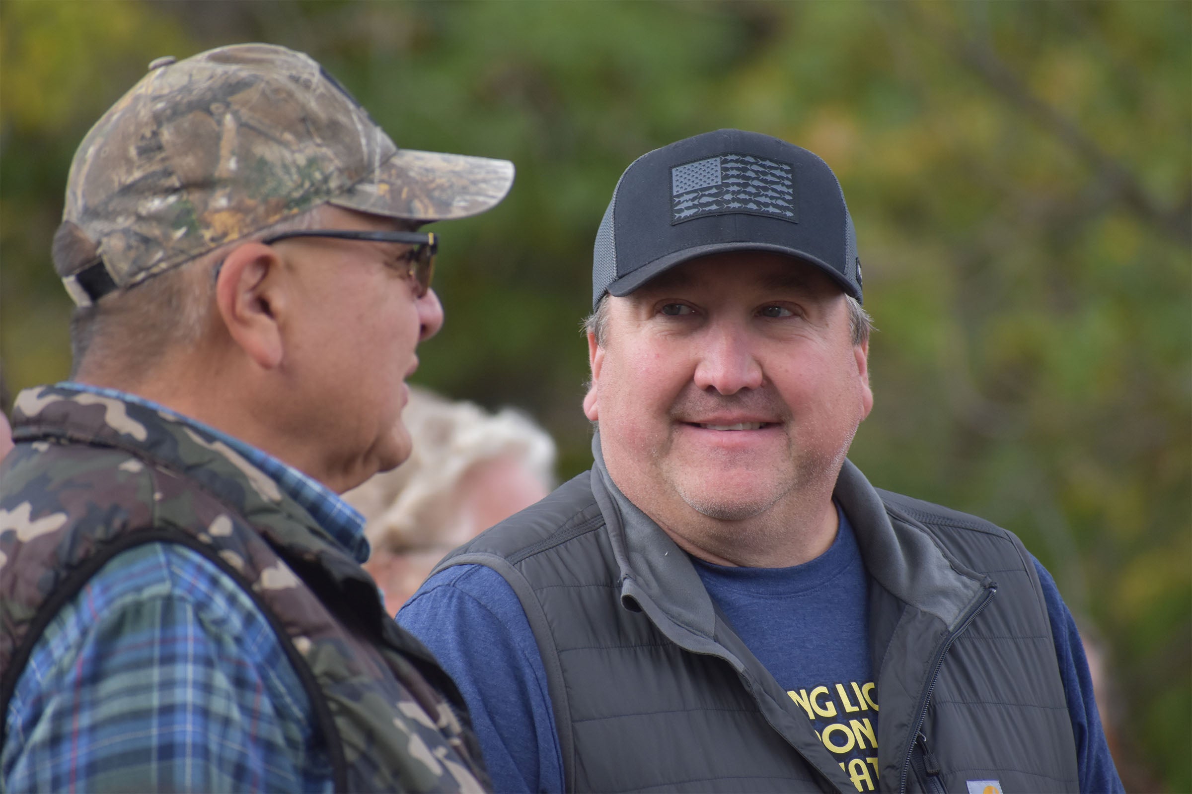 Two men outdoors talk to each other; one wears a camouflage cap and glasses, the other a black cap and gray vest. Trees and a blurred figure are in the background.