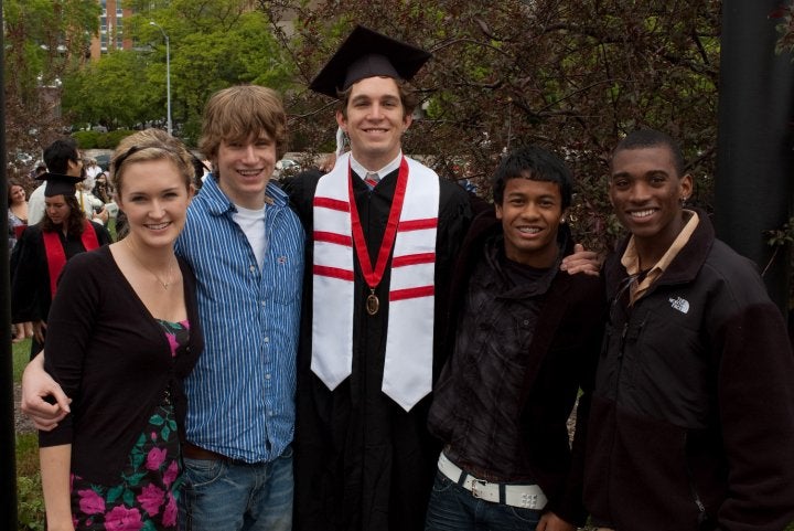 Five young adults stand outdoors, with one wearing a graduation cap and gown. The group is smiling, and trees and people are visible in the background.